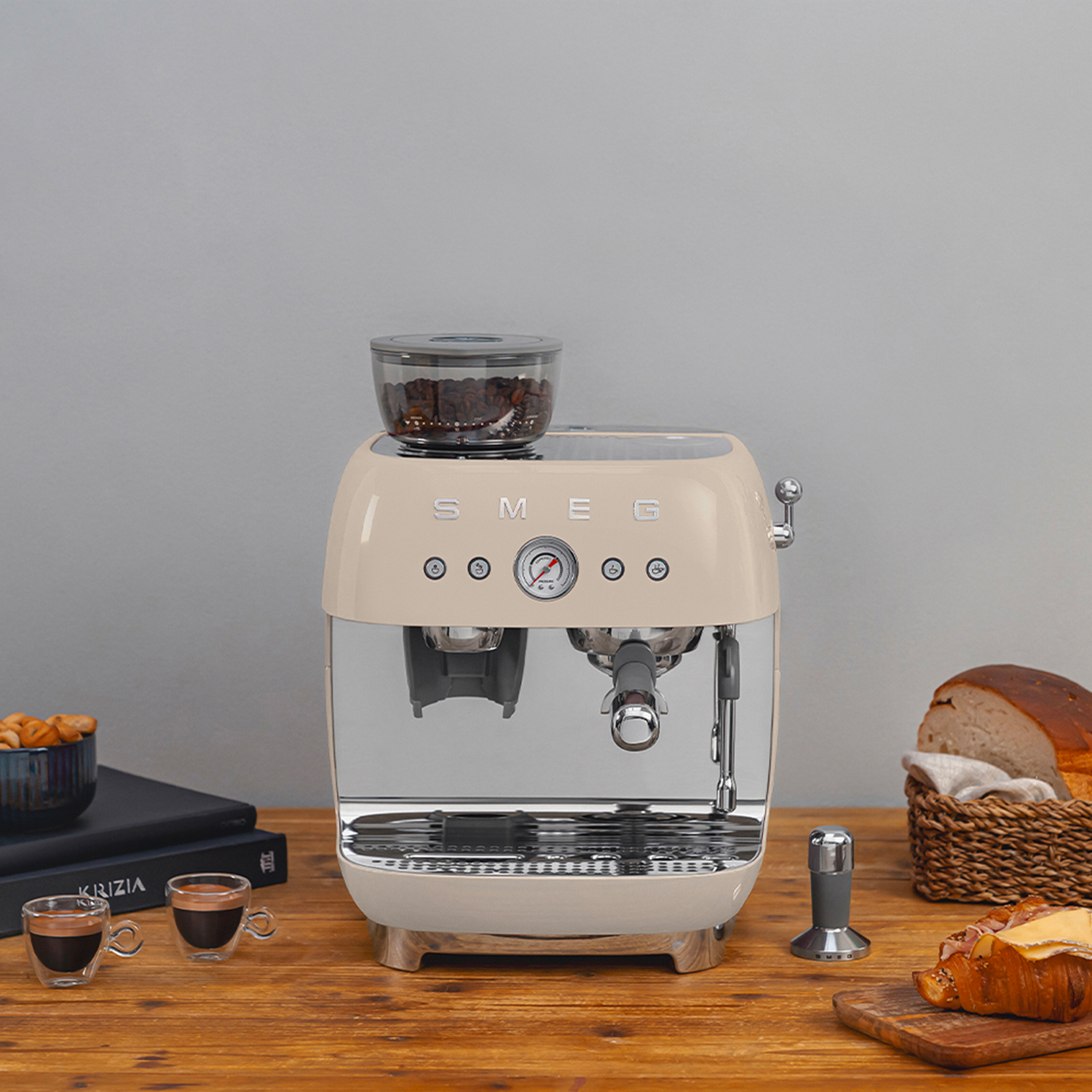 Beżowa ekspres Smeg z zasobnikiem na ziarna, dwoma filiżankami, chlebem i rogalikiem.