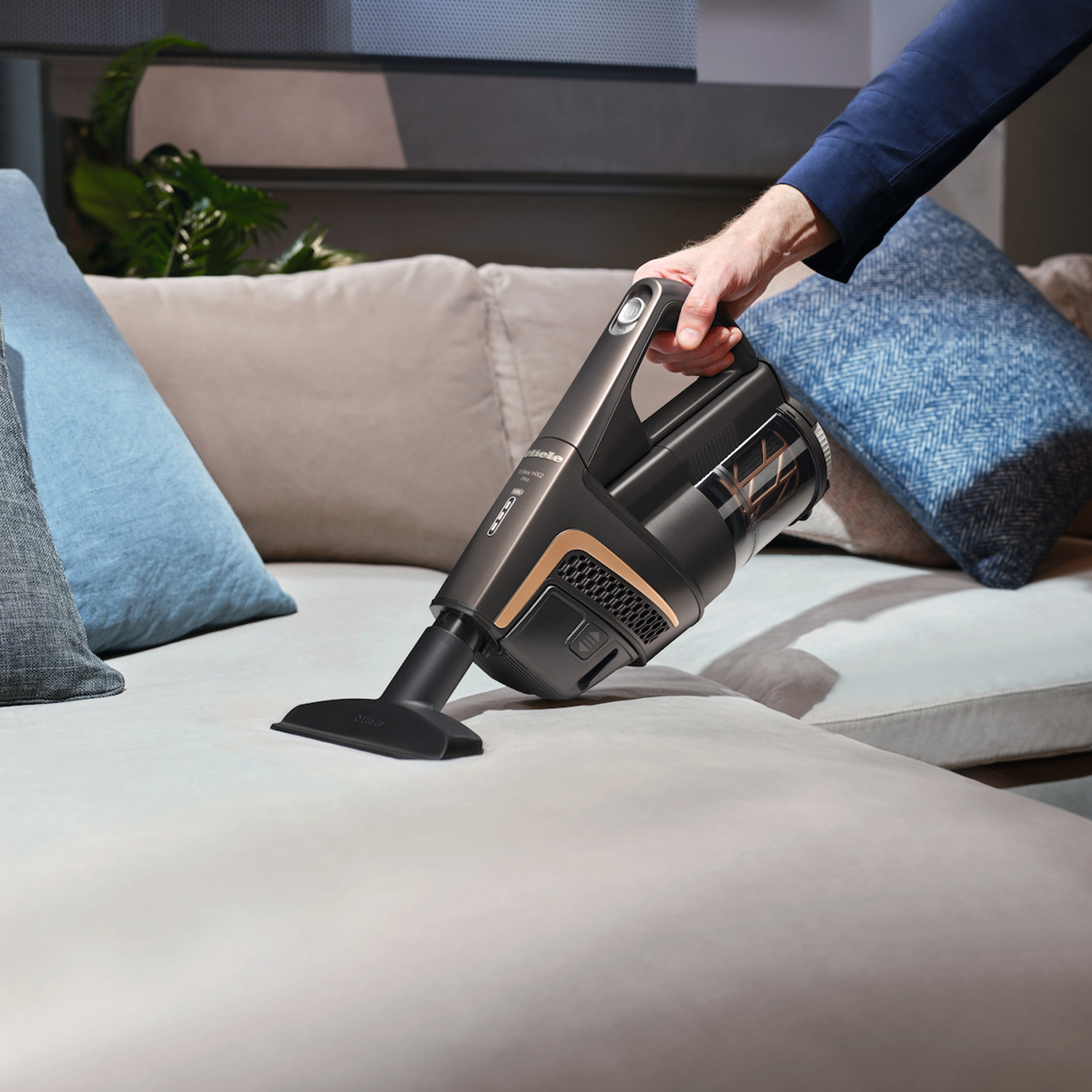 Eine Hand hält einen dunkelgrauen Akku-Staubsauger und reinigt ein hellgraues Sofa.