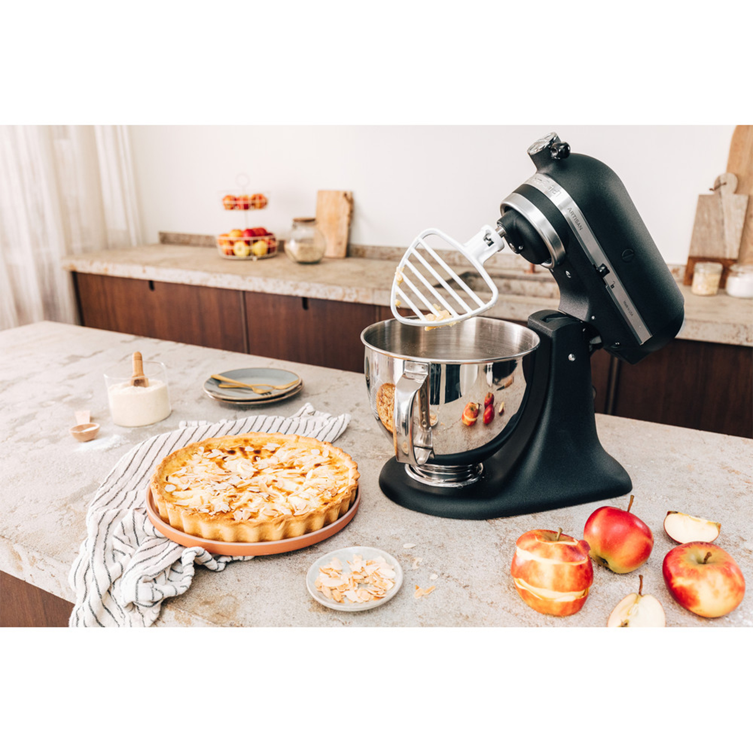 Küchenmaschine mit Schüssel, Apfelkuchen, Äpfeln und Mandelsplittern auf einer Arbeitsplatte.