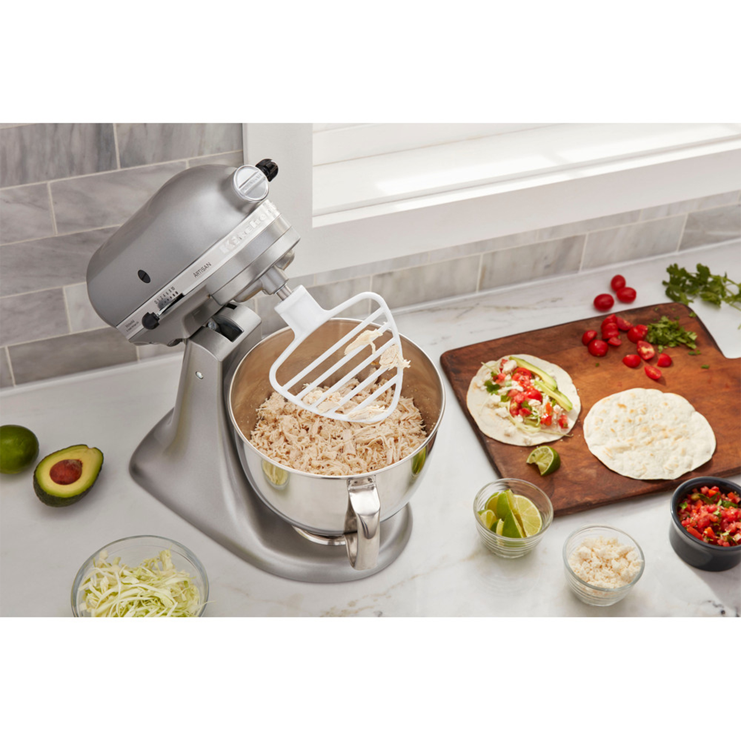 Silberner Standmixer mit Schüssel voller zerrupfter Hähnchenbrust. Tortillas und Zutaten daneben.