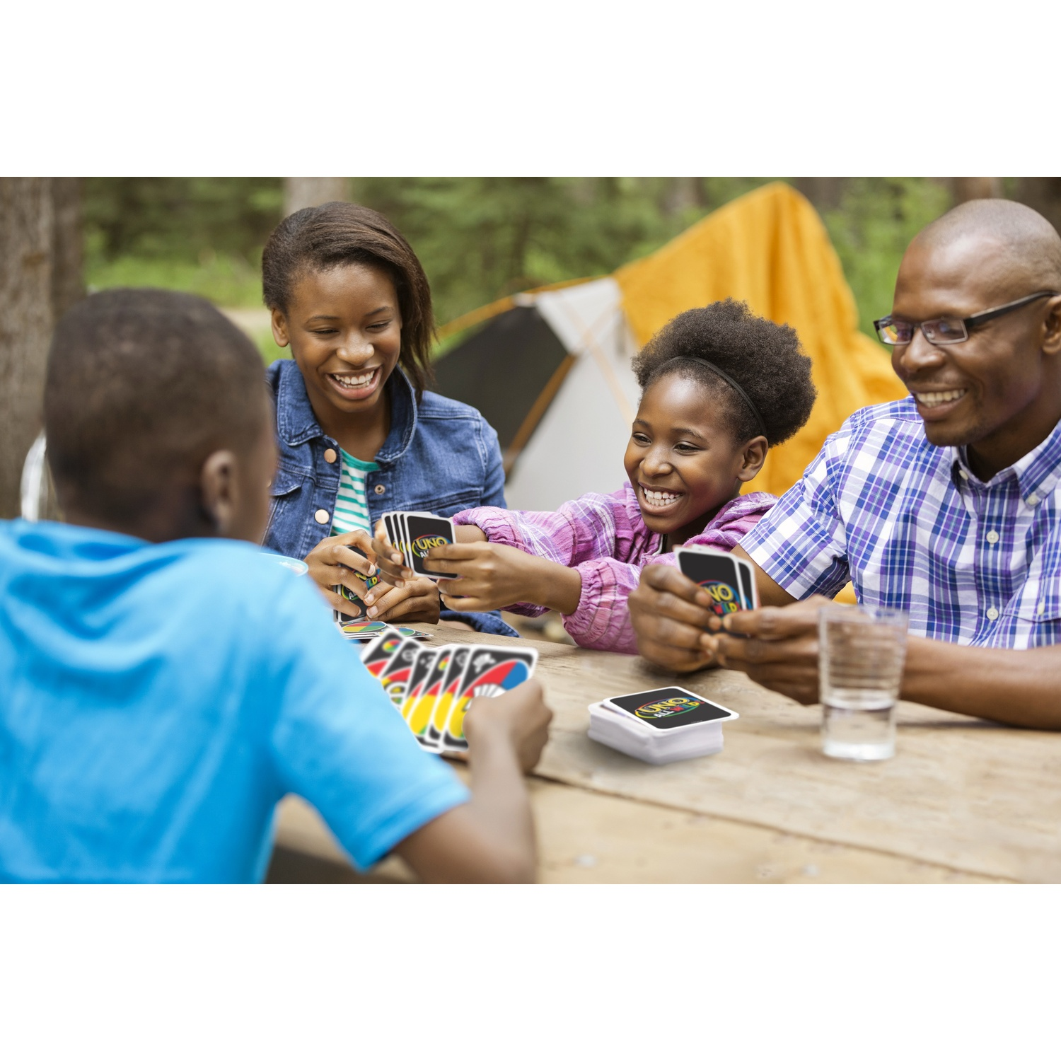 Une famille joue à UNO en plein air à une table en bois avec une tente en arrière-plan.