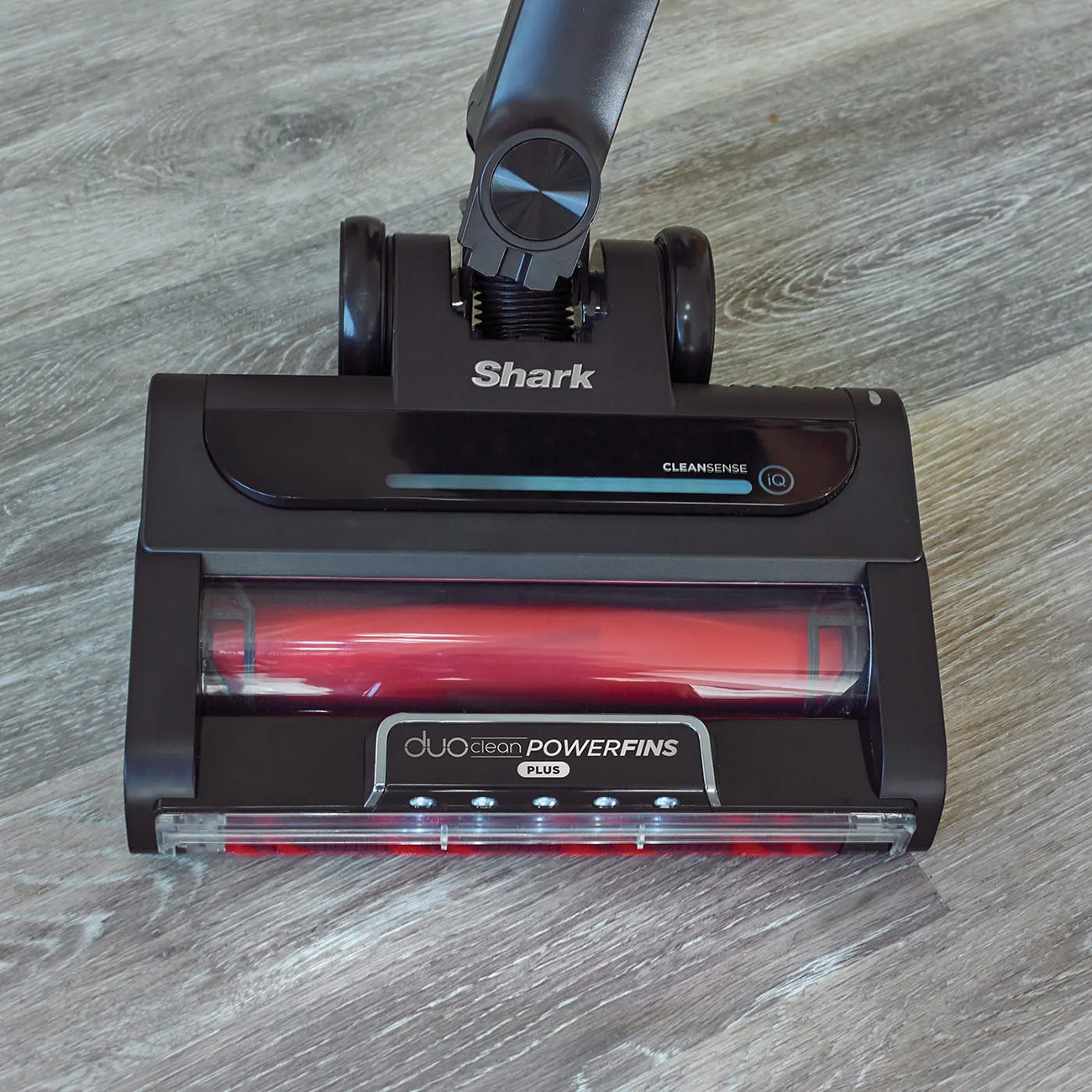 Un cabezal de aspiradora Shark negro con cepillo rojo sobre suelo de madera gris.
