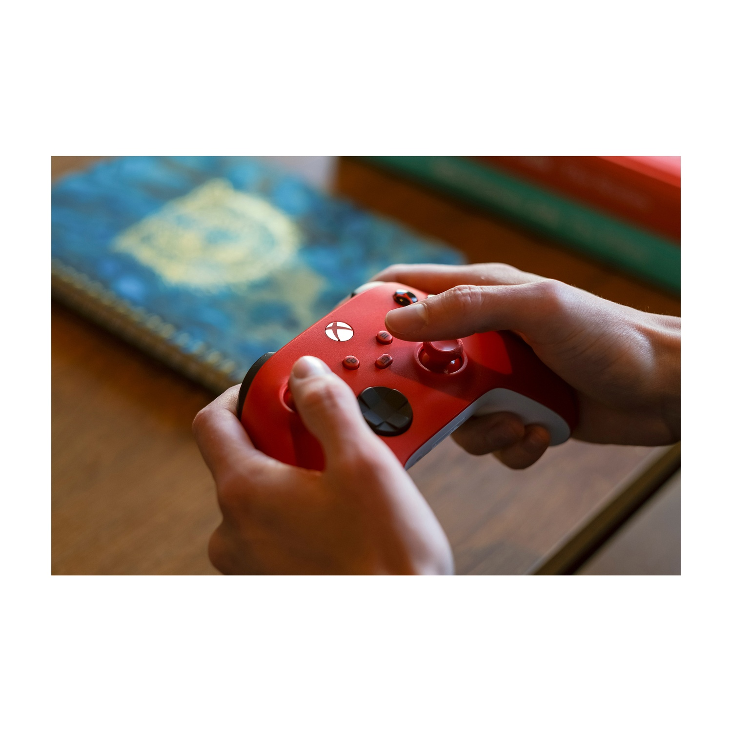 Handen houden een rode Xbox controller vast op een houten tafel met boeken op achtergrond.