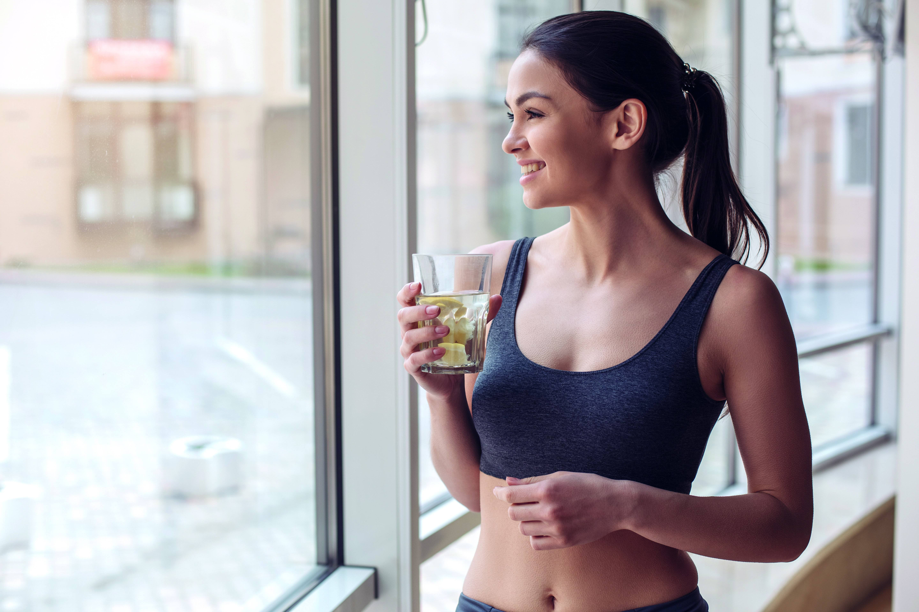 Kobieta trzyma szklankę wody z cytryną i patrzy przez okno.