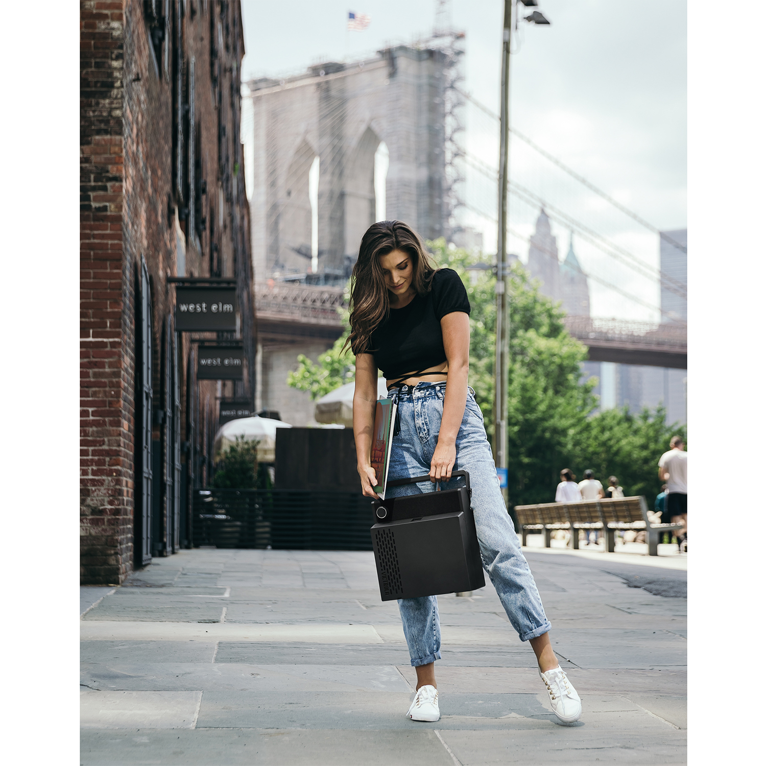 Una mujer con jeans y top negro lleva un maletín negro. Puente de Brooklyn al fondo.
