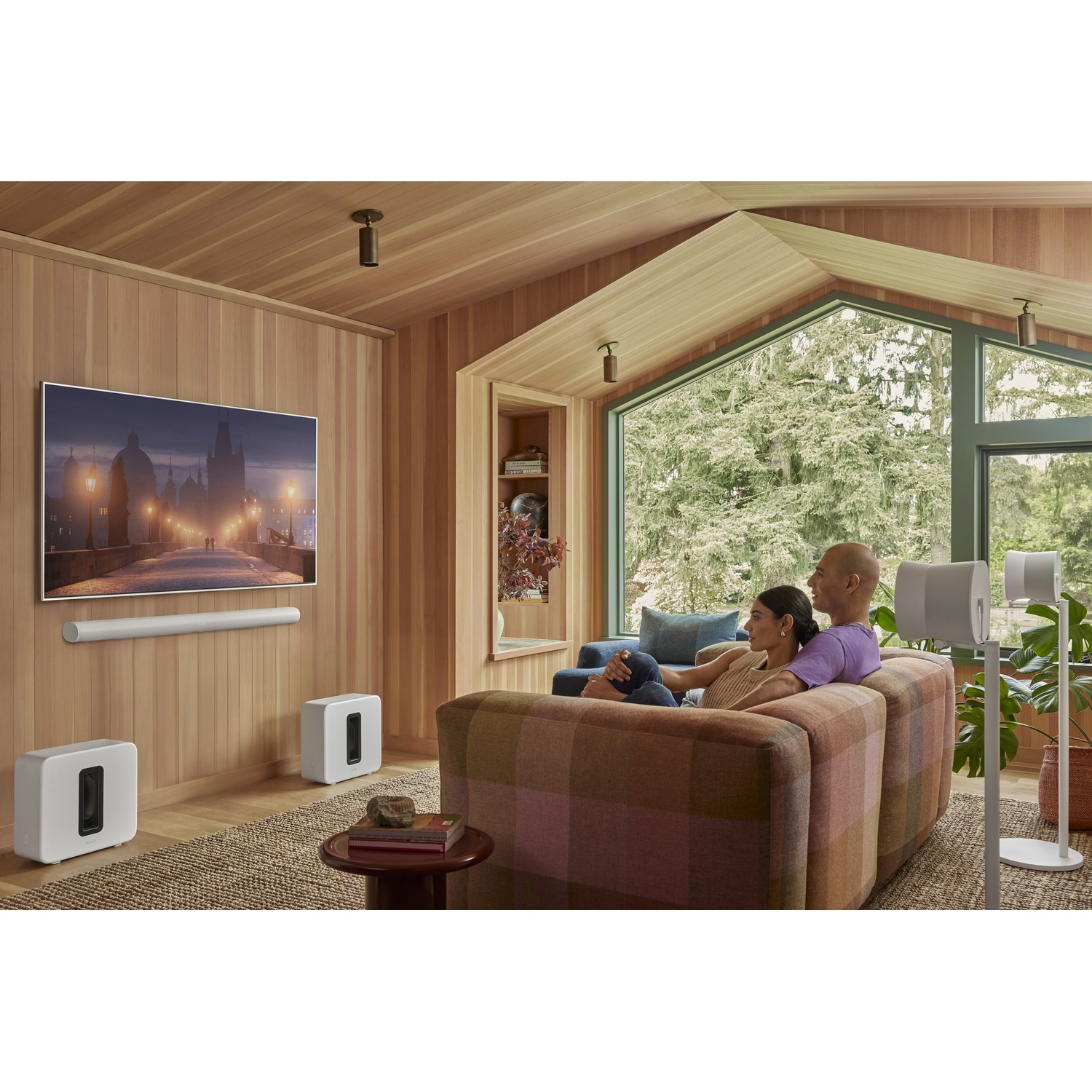 Para na kanapie ogląda telewizję. Białe głośniki i soundbar w pokoju z drewnianymi panelami.