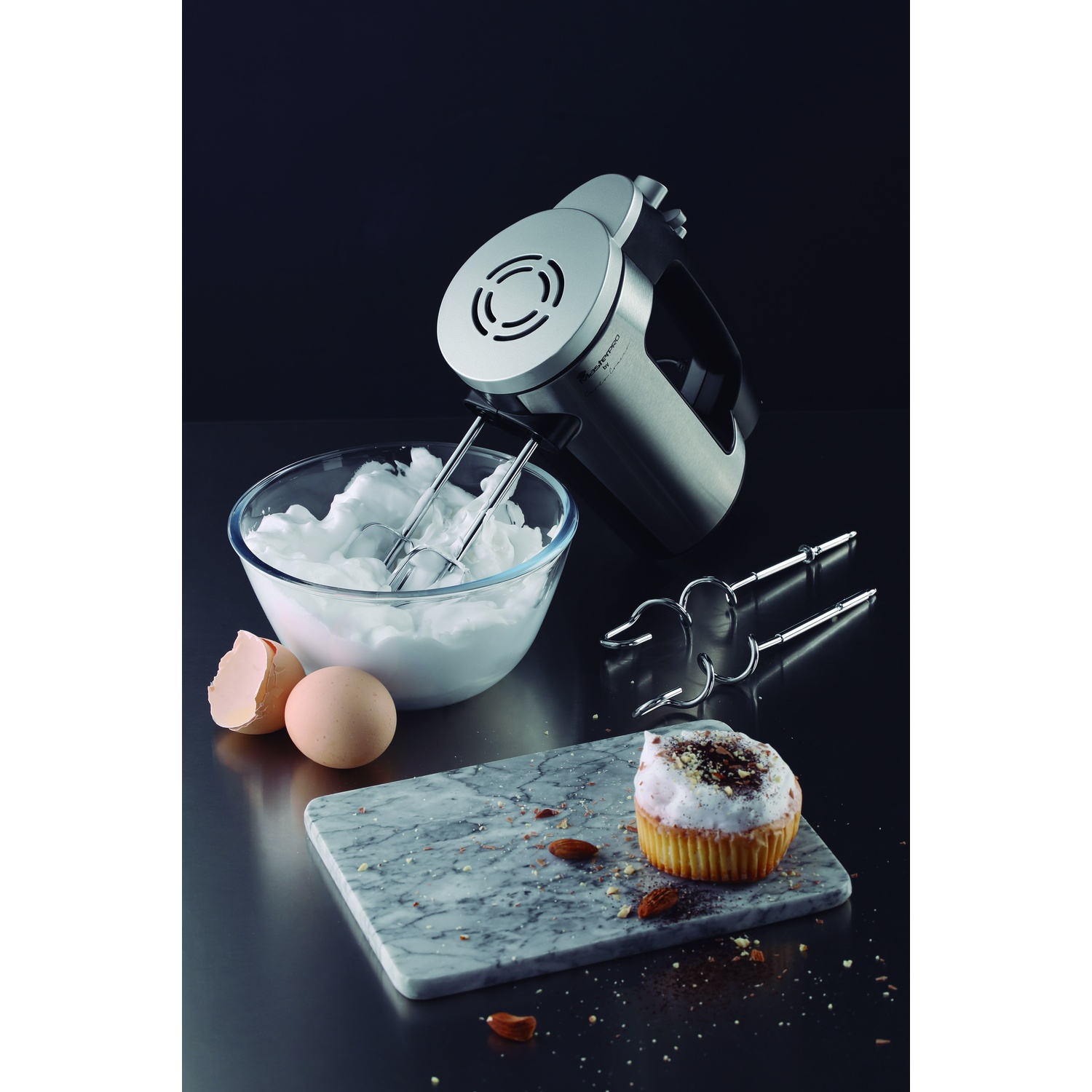 Una batidora de mano está junto a un bol de merengue, huevos y un cupcake.