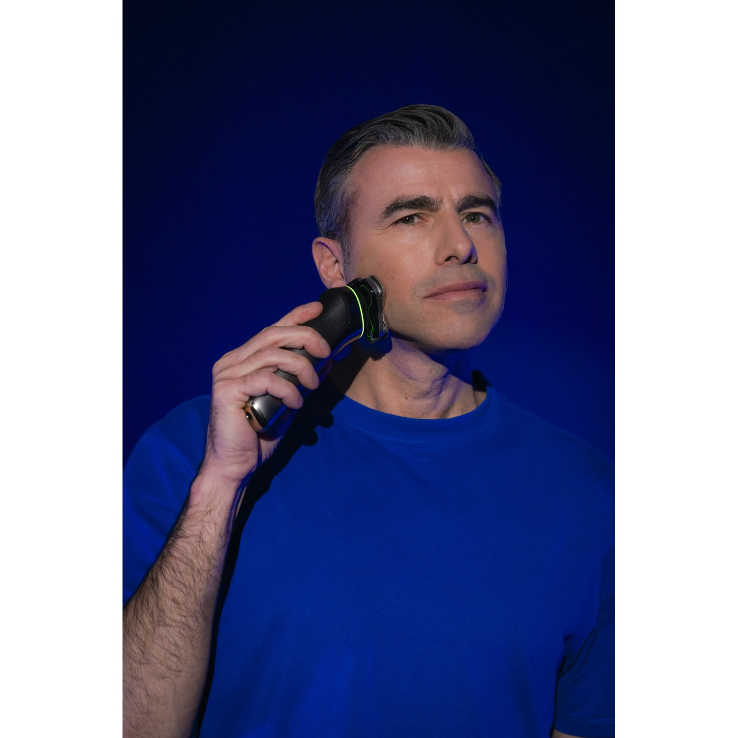 Hombre con afeitadora eléctrica arreglándose la cara, con camisa azul y fondo azul.