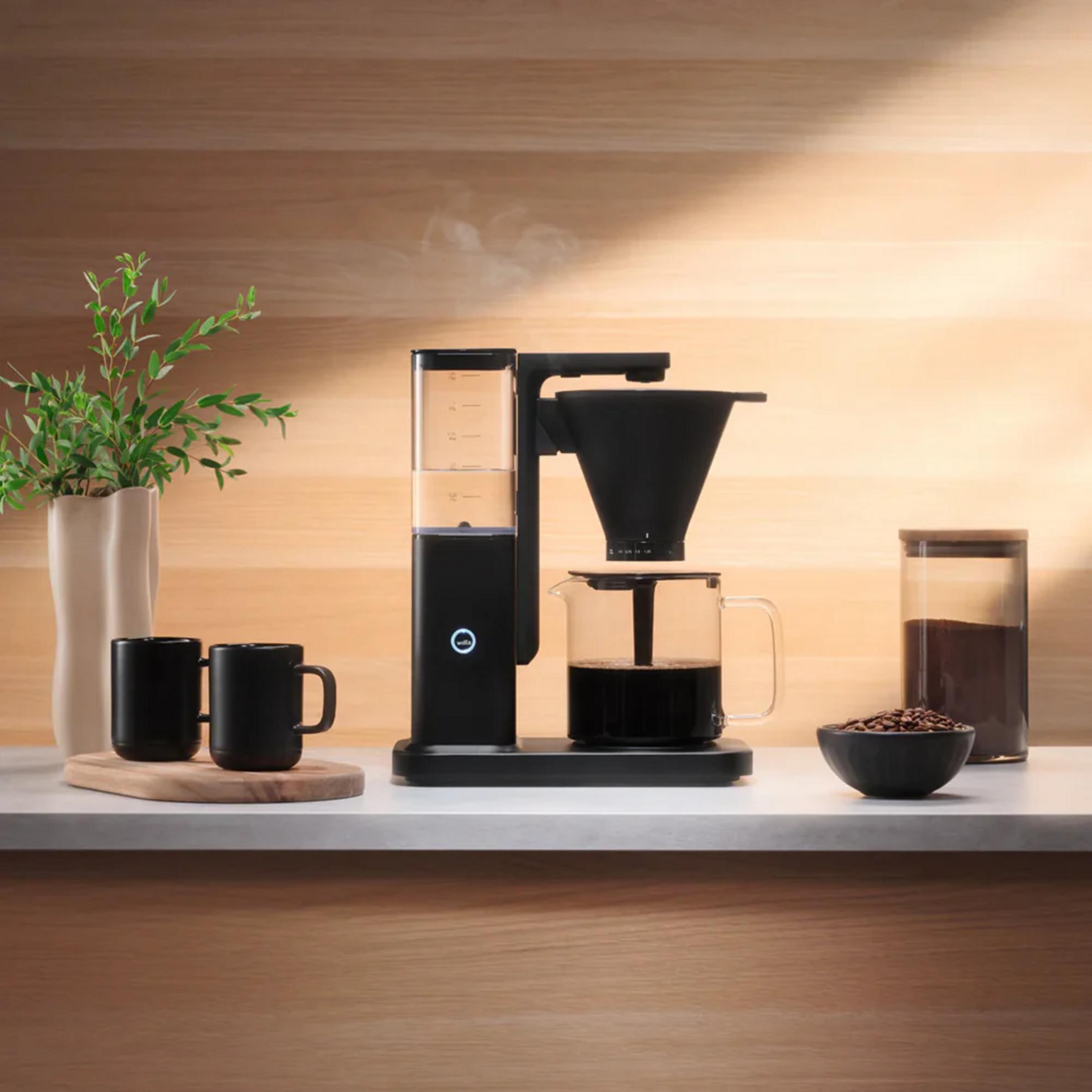 Macchina da caffè con caraffa, tazze, chicchi e piante su un piano di lavoro.