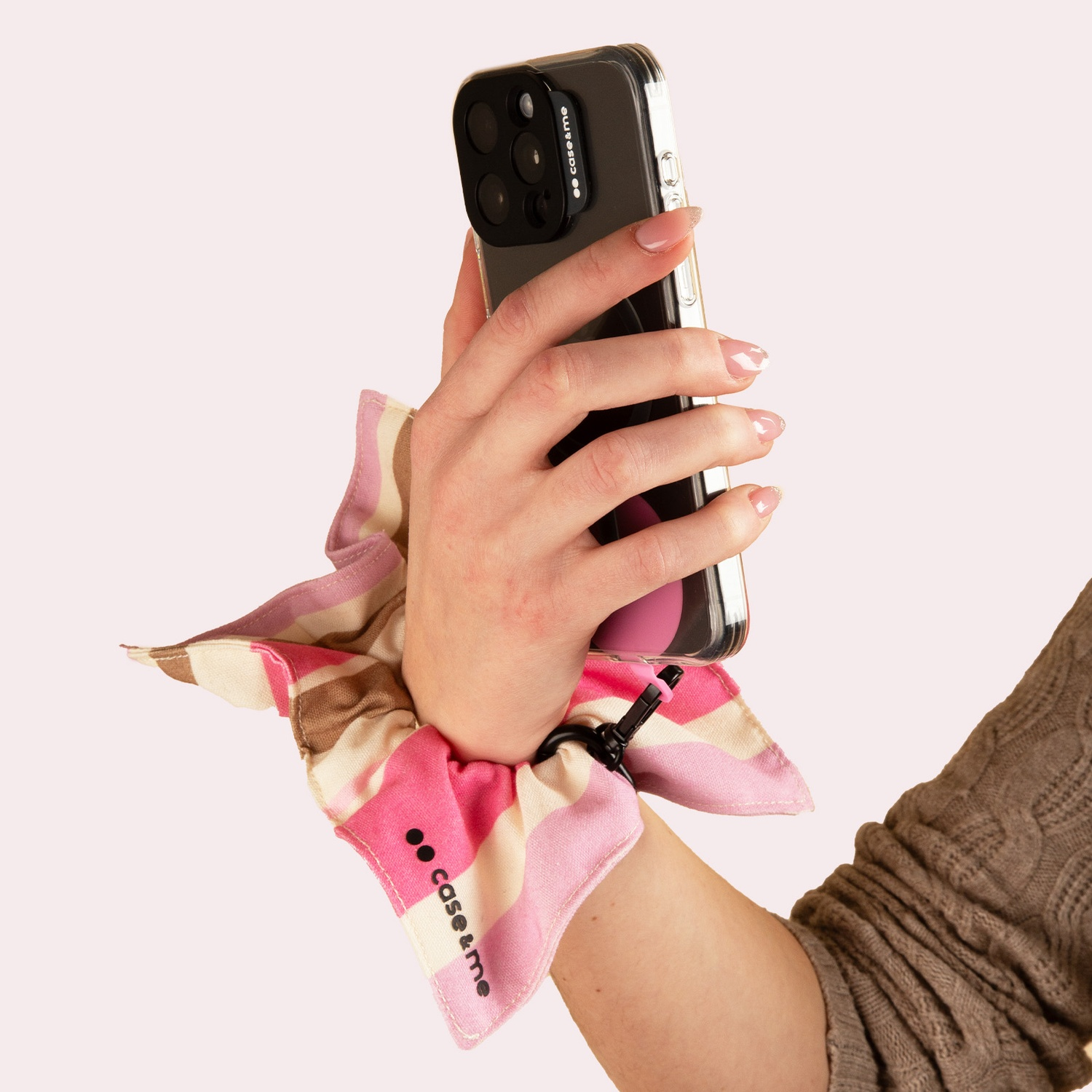 Mano tiene smartphone con cinturino da polso a righe rosa e bianche. Sfondo neutro.
