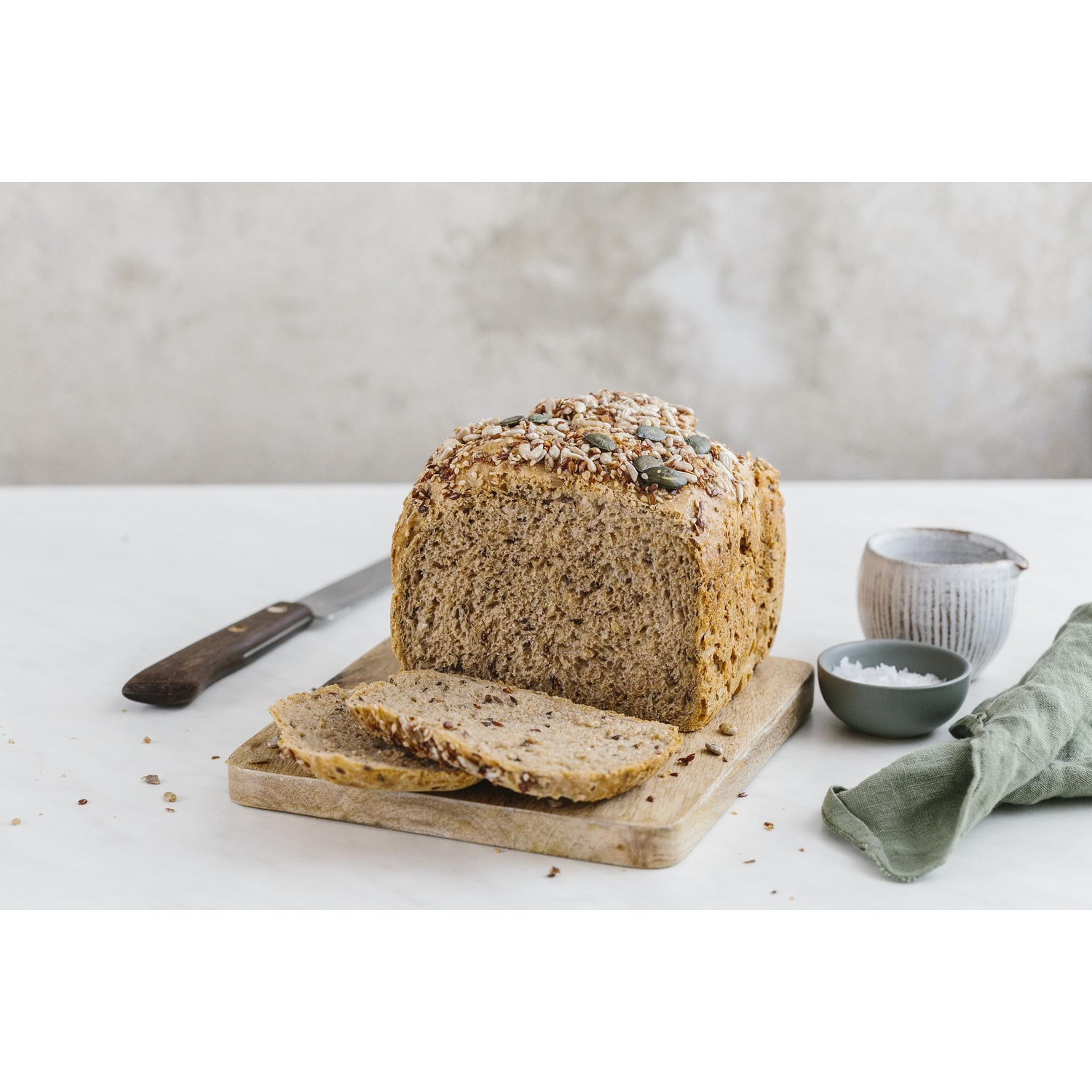 Een brood met zaden en twee plakken op een houten plank, mes, zout en doek ernaast.