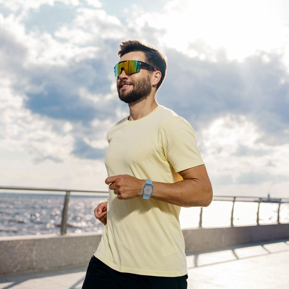 Mann mit Bart und Sonnenbrille joggt in einem gelben Hemd am Wasser.
