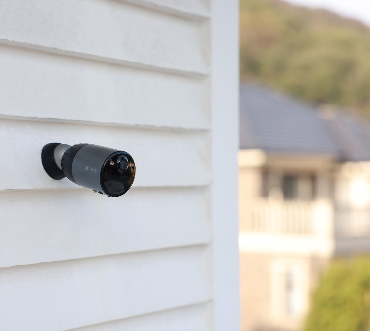 Ciemna kamera zamontowana na białej ścianie. Niewyraźne domy w tle.