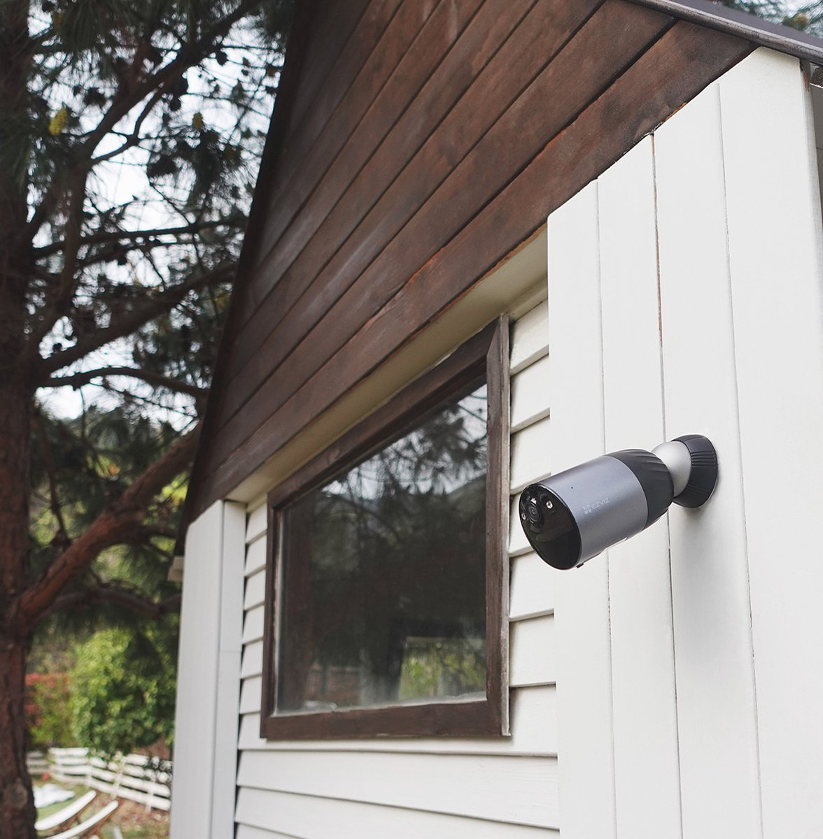 Szara kamera bezpieczeństwa zamontowana na białej ścianie obok okna drewnianego domu.