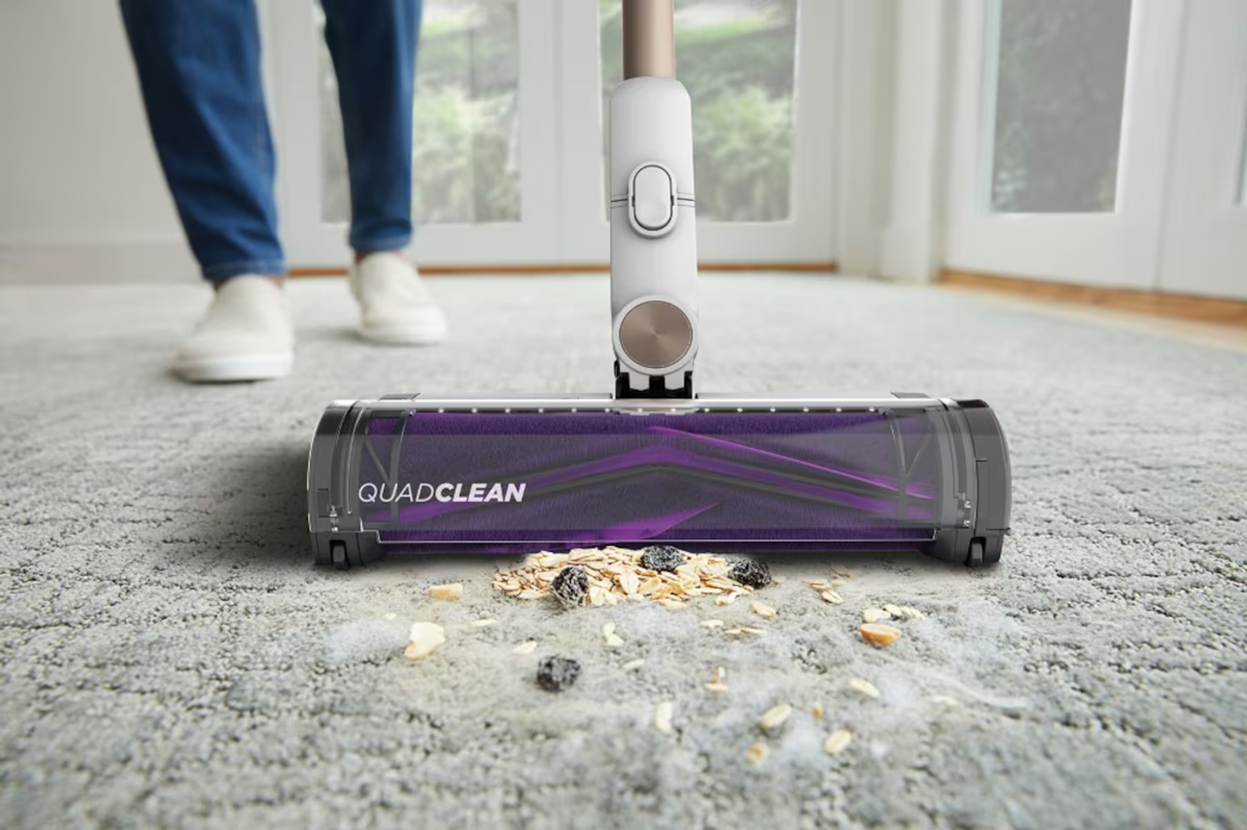 A vacuum cleaner head with purple brushes picks up crumbs from a carpet.