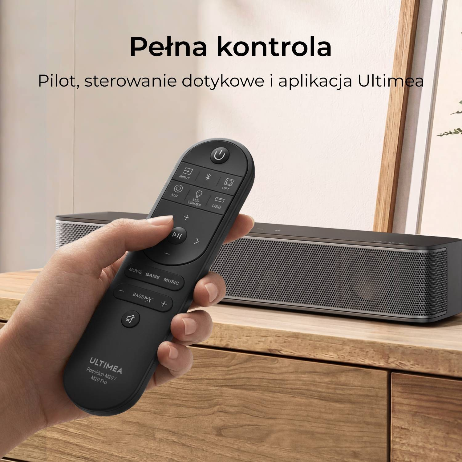 Dłoń trzyma czarny pilot przed czarną listwą głośnikową na drewnianej szafce.