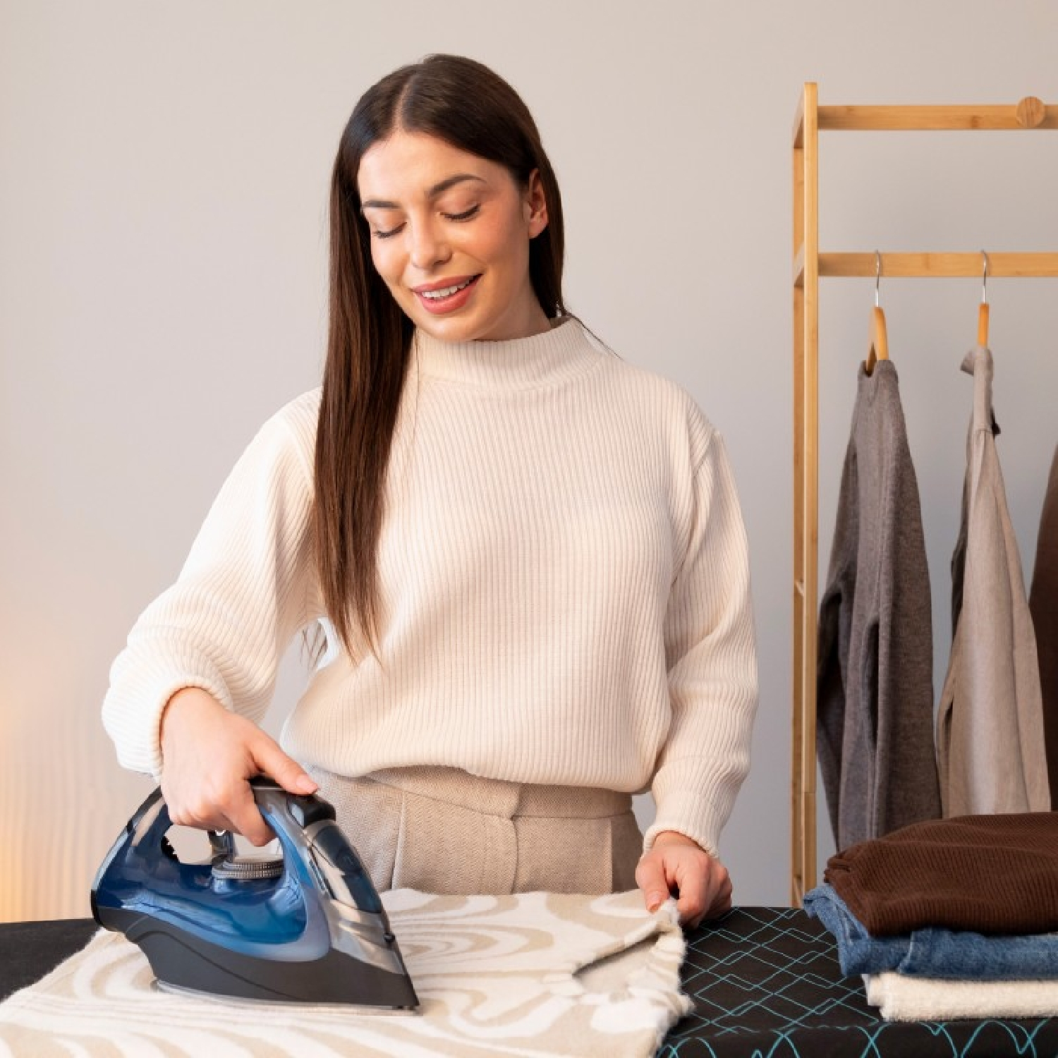 A woman irons clothes on an ironing board, with clothes racks in the background.