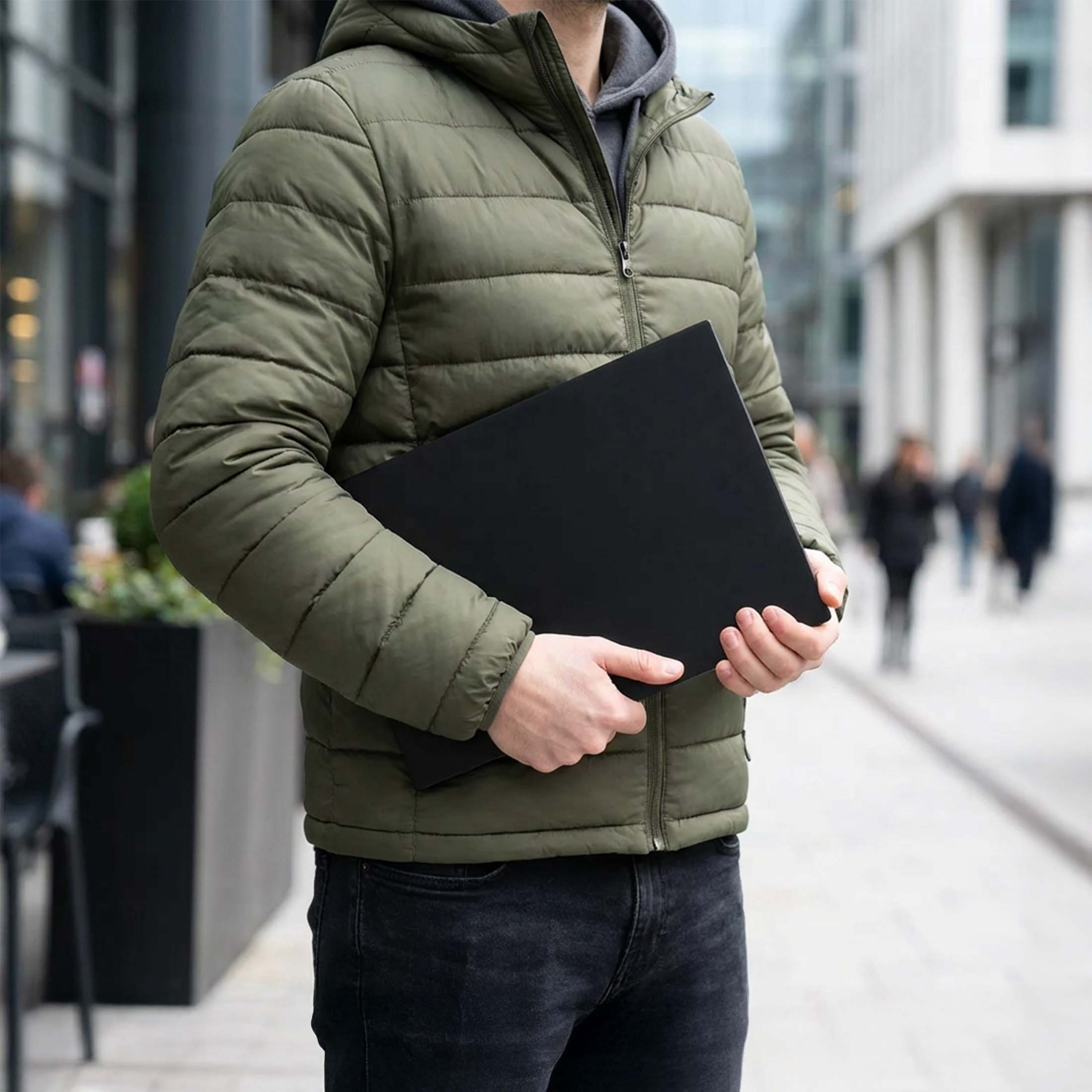 Mann in grüner Jacke hält schwarzen Laptop auf der Straße.