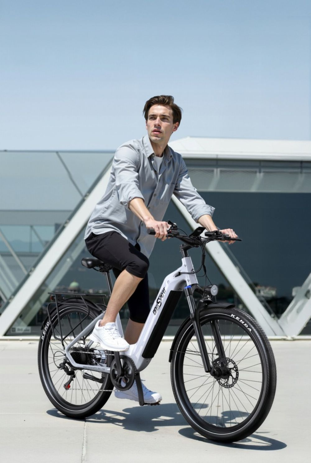 Un hombre con camisa gris monta una bicicleta eléctrica blanca frente a un edificio moderno.