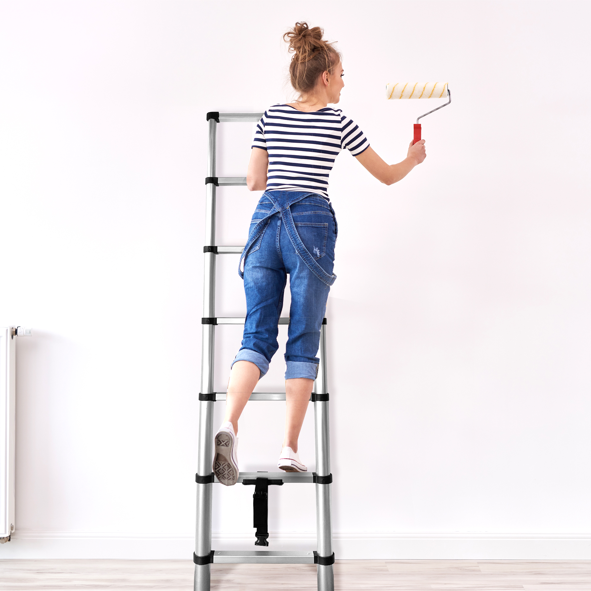 Mujer en escalera pintando pared con rodillo. Lleva overol de mezclilla y camisa.