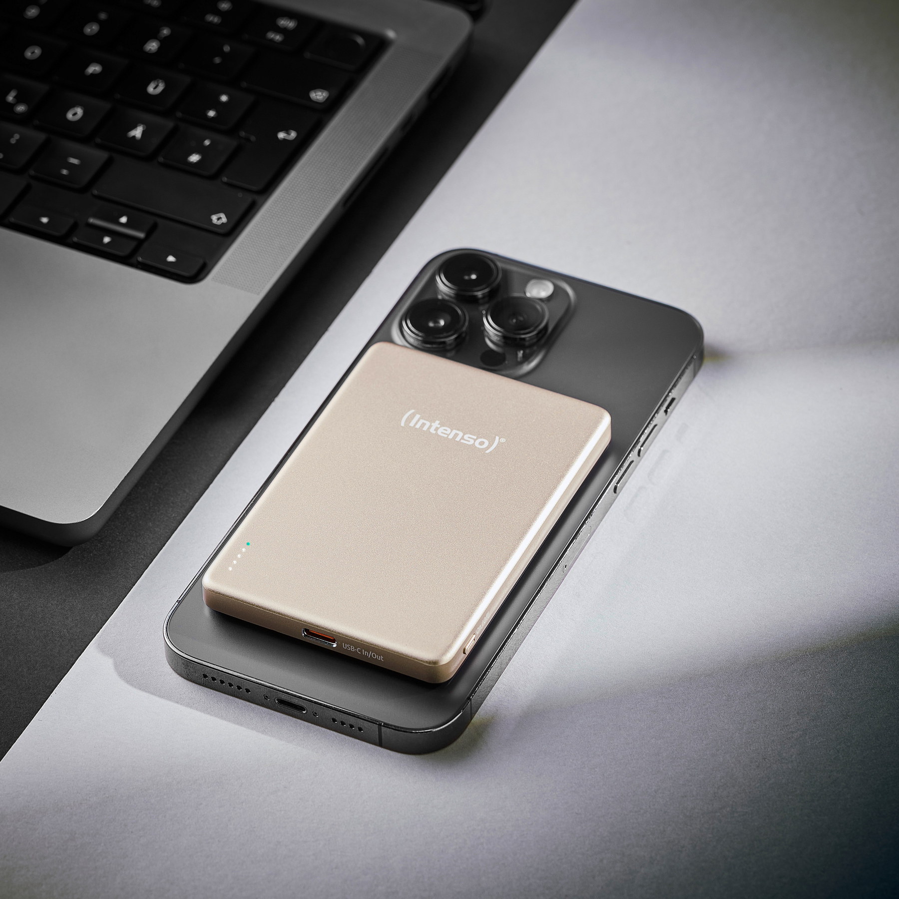 Un power bank Intenso dorato poggia su uno smartphone scuro accanto a un laptop.