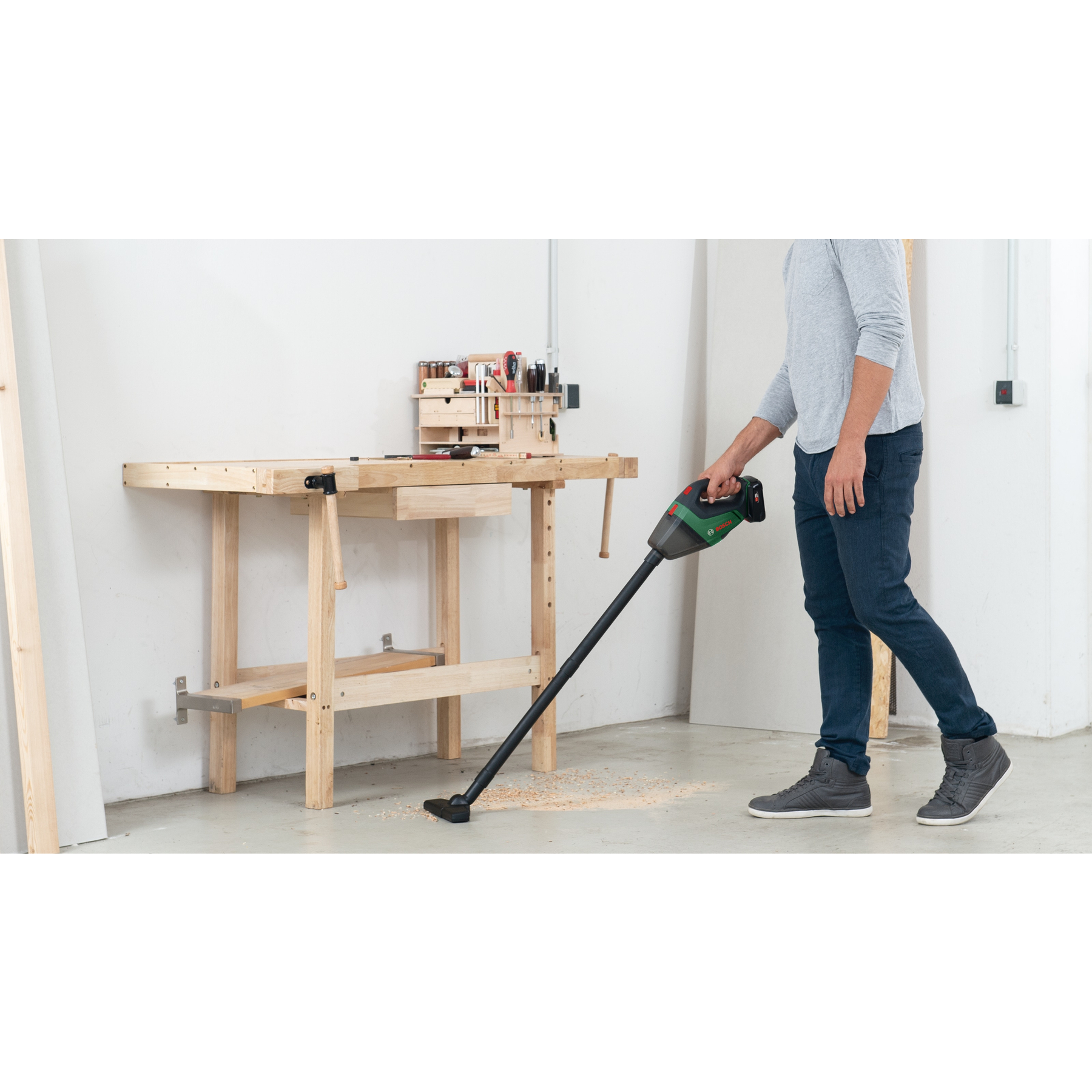 Hombre aspira virutas de madera con aspiradora verde-negra cerca de banco.