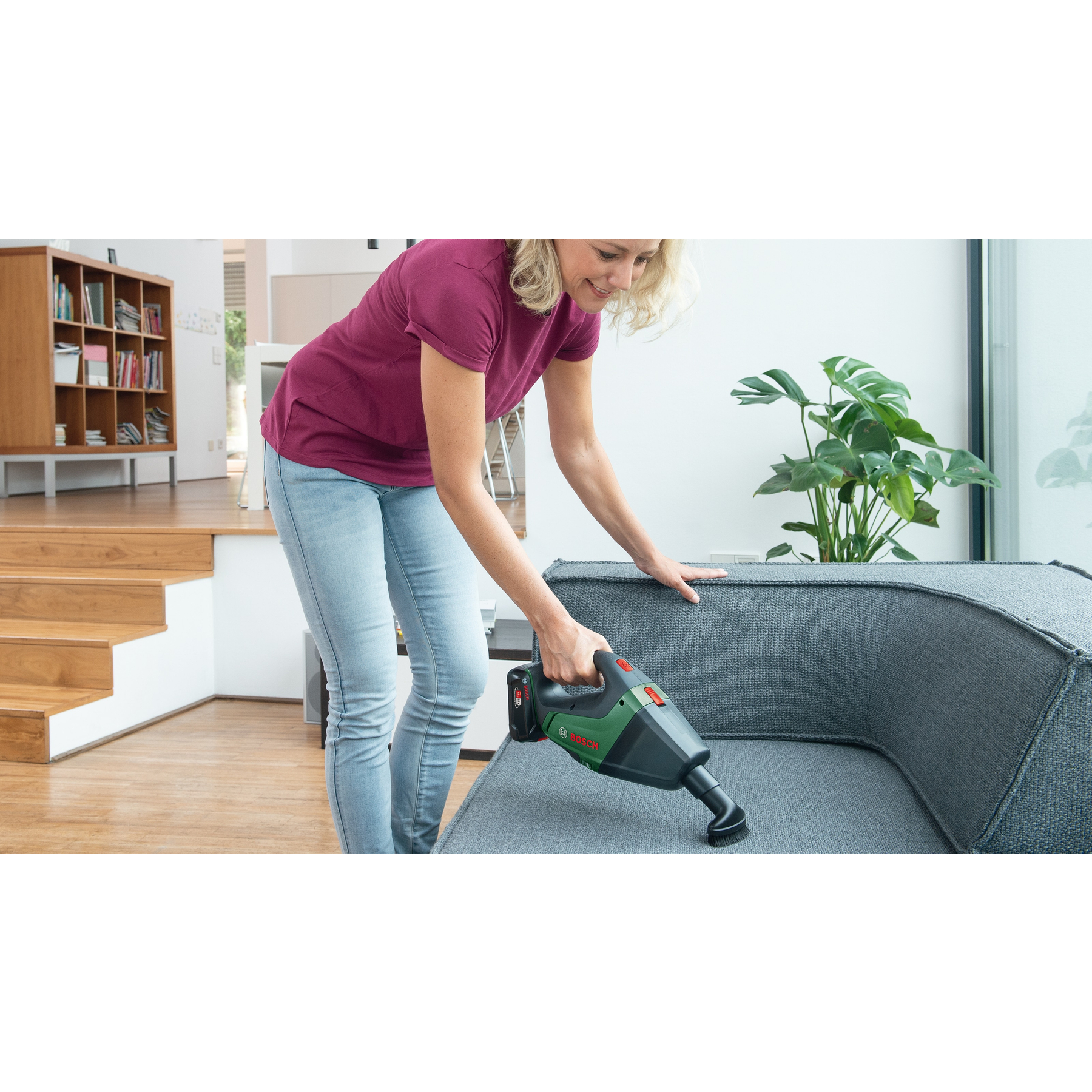 Mujer limpia sofá con aspiradora de mano. Escaleras de madera y estantería atrás.