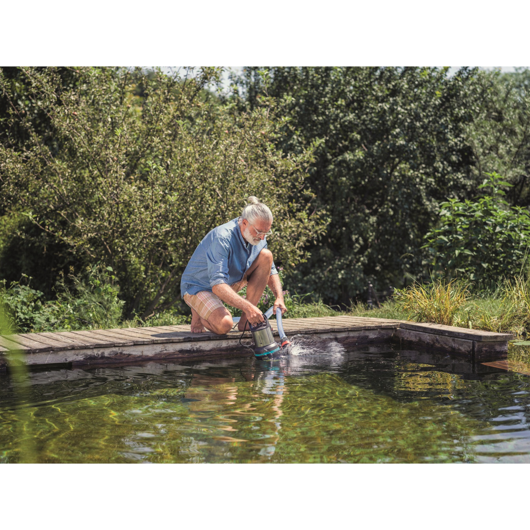 Mann mit grauem Haar reinigt Teich mit Pumpe auf Holzsteg.