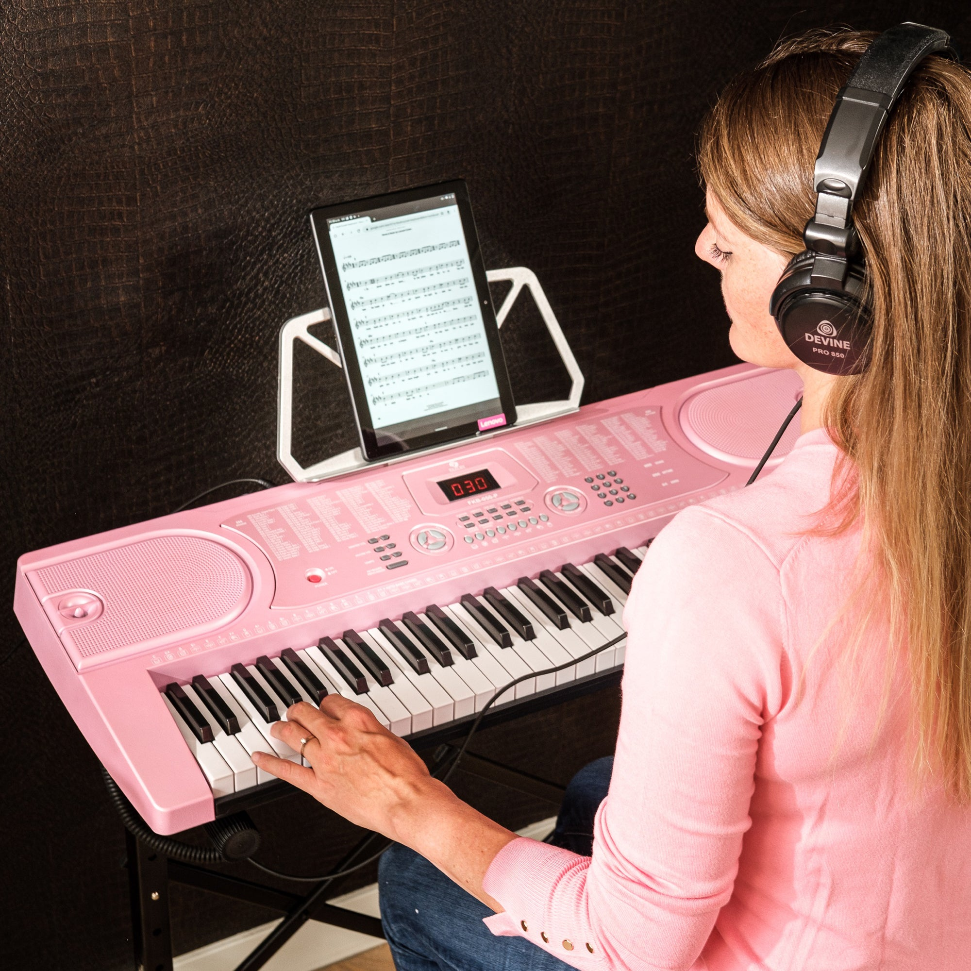 Een vrouw met koptelefoon speelt op een roze keyboard, met bladmuziek op een tablet.