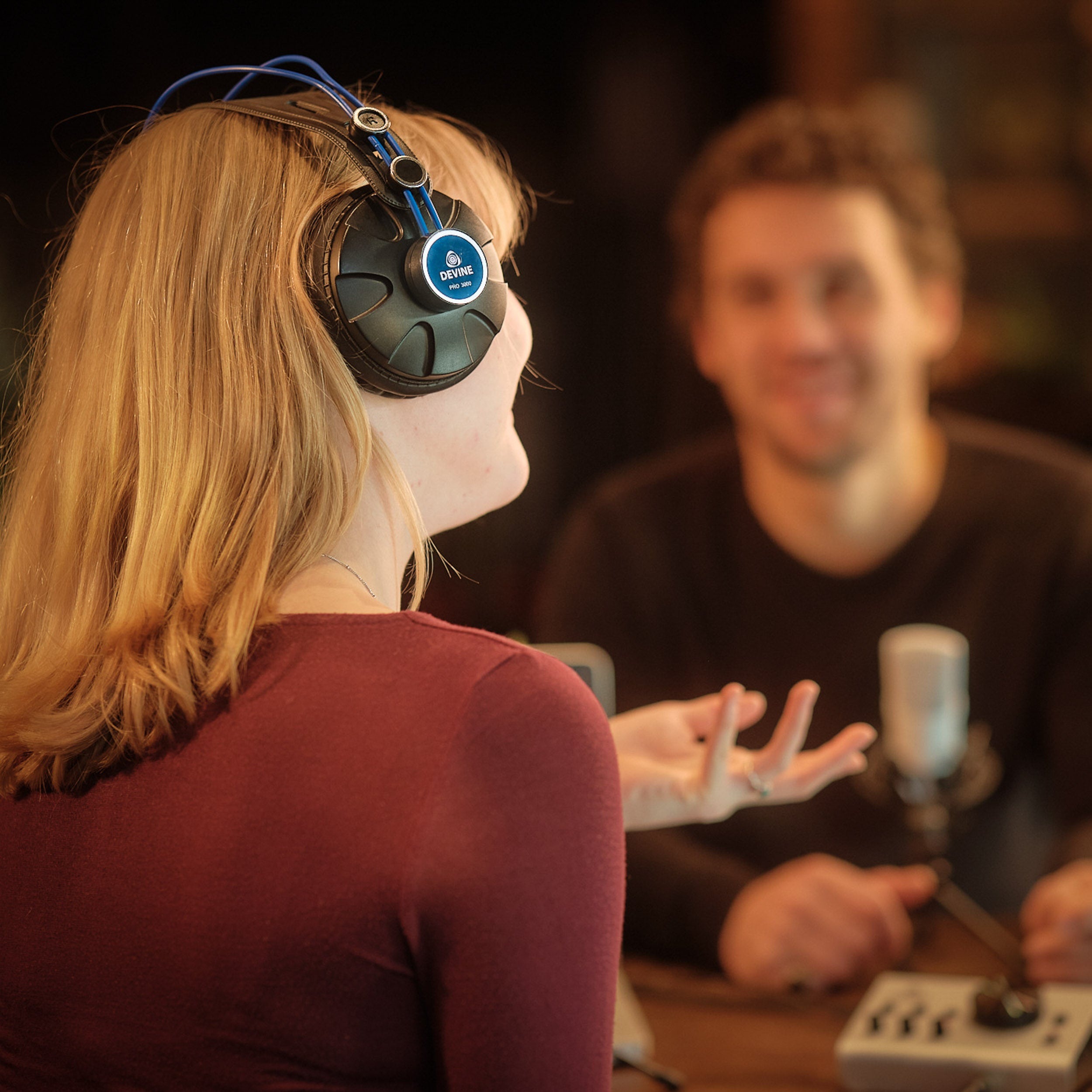 Vrouw met koptelefoon spreekt microfoon, man lacht op achtergrond.