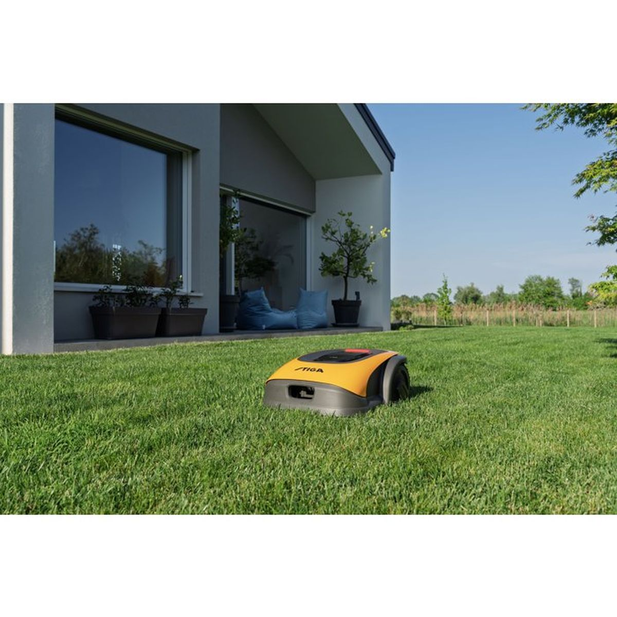 Un robot tosaerba giallo e grigio si trova su un prato verde davanti a una casa moderna.