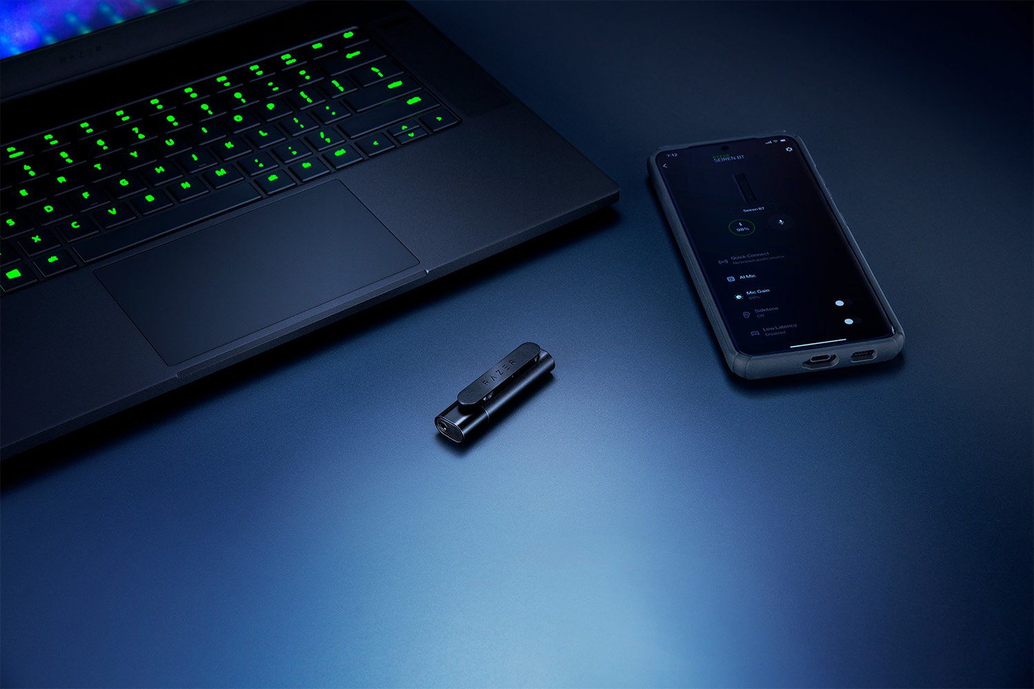 A black laptop with green keys, a dongle, and a smartphone rest on a blue surface.