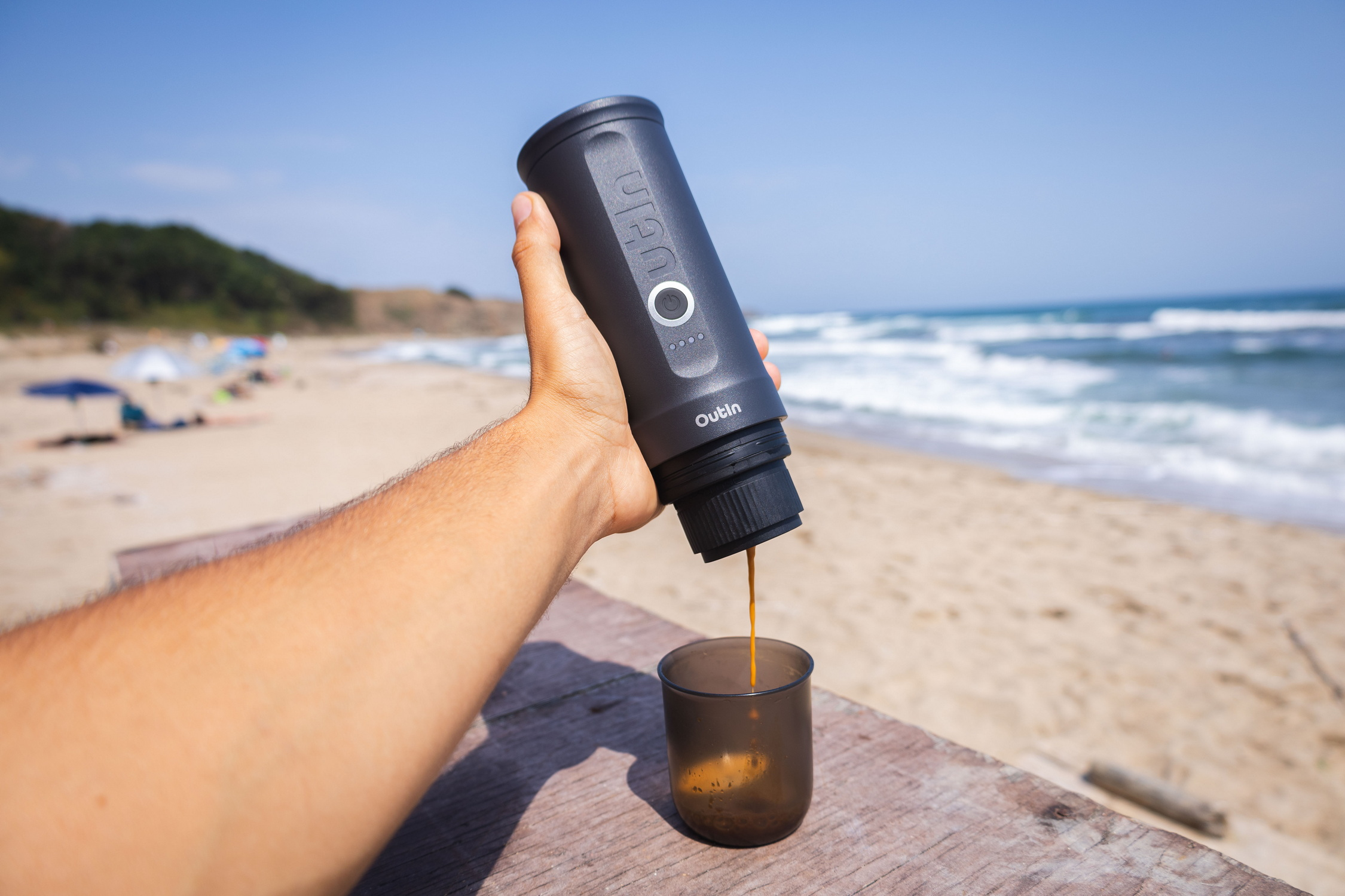 Een hand houdt een koffiezetapparaat vast dat koffie in een glas op een strand schenkt.