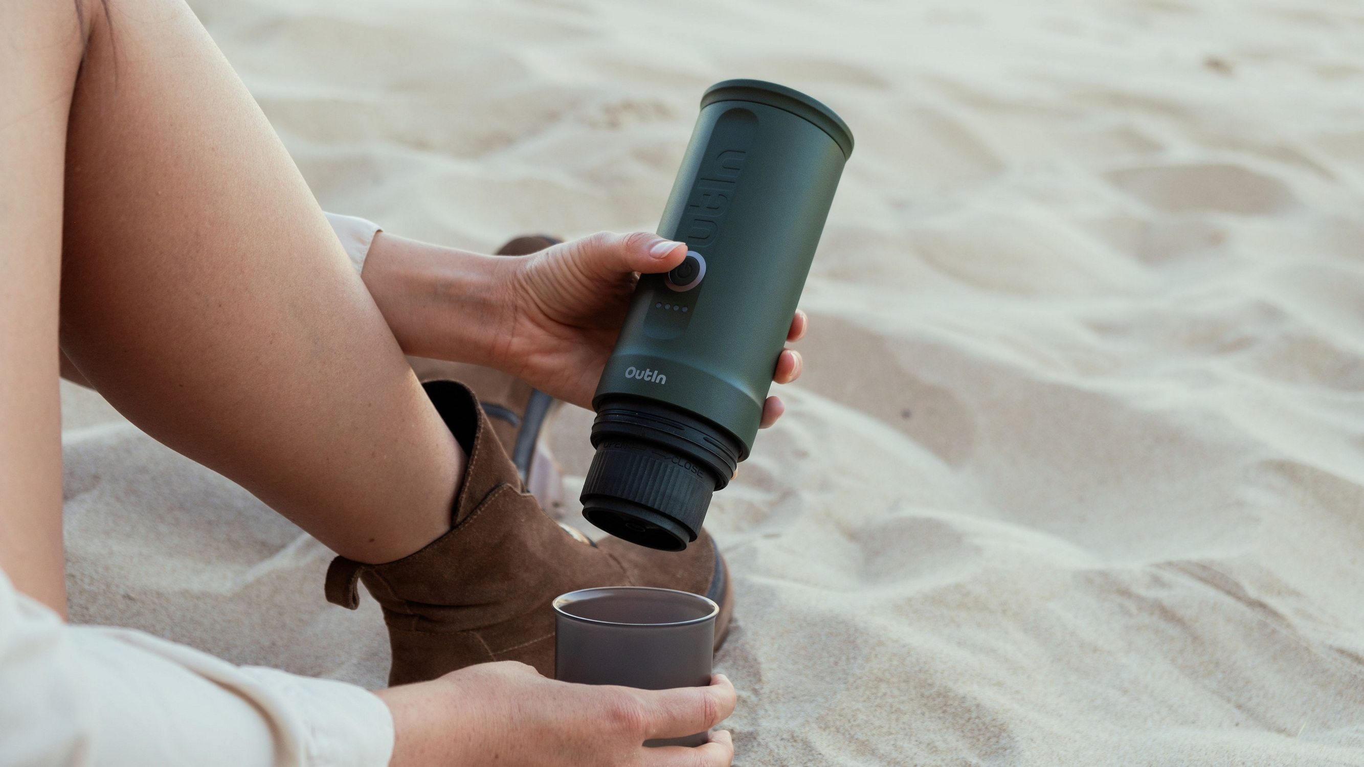 Een hand houdt een groene waterfles boven een beker op het strand.