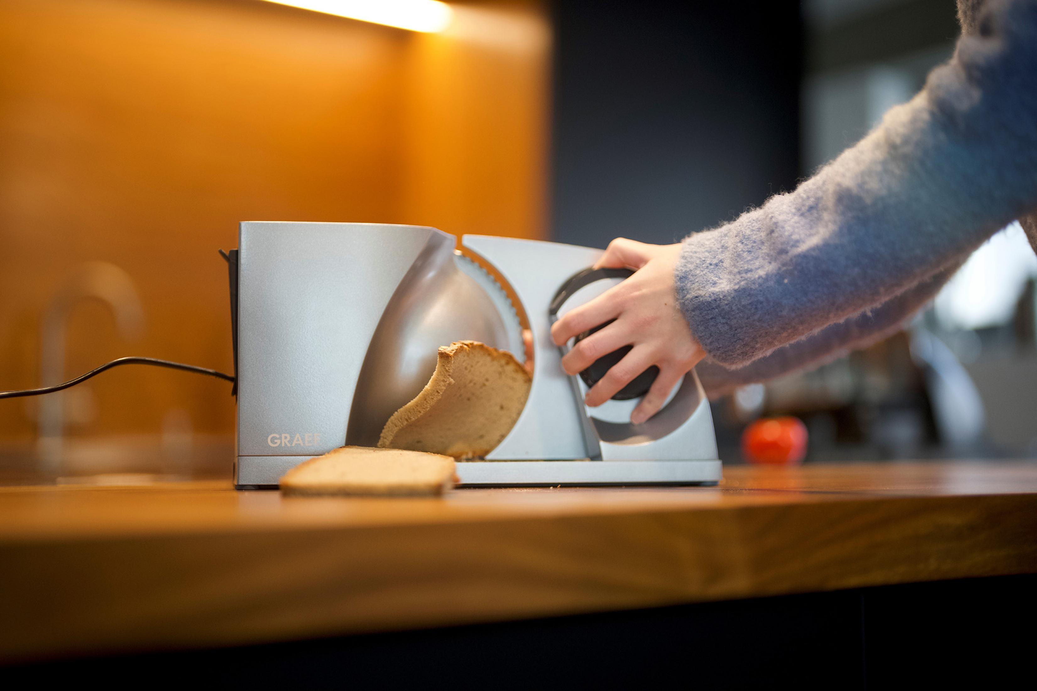 Eine Person schneidet Brot mit einem silbernen elektrischen Brotschneider in einer Küche.