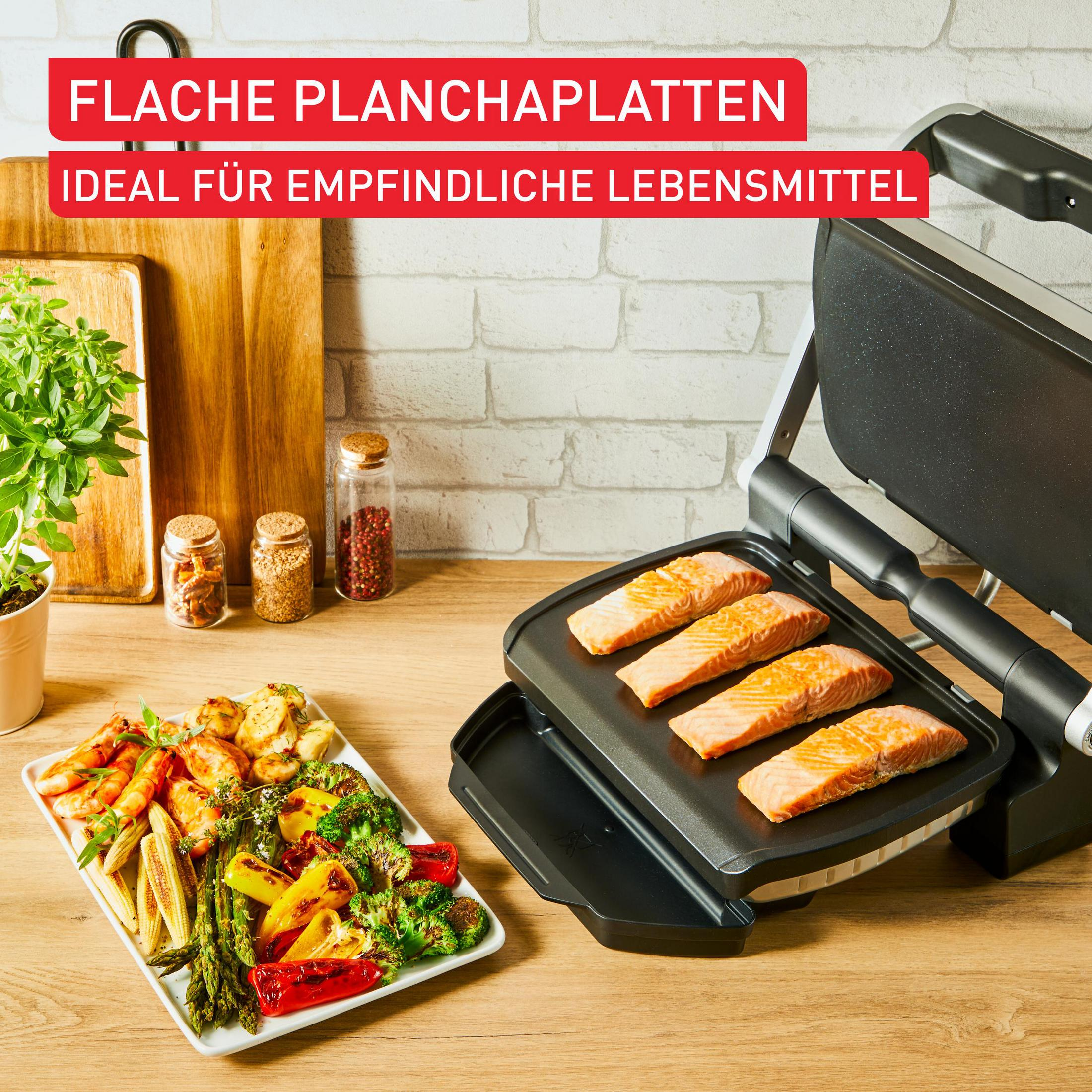 Grill mit Lachs und Gemüse auf Holztisch, weiße Ziegelwand im Hintergrund.