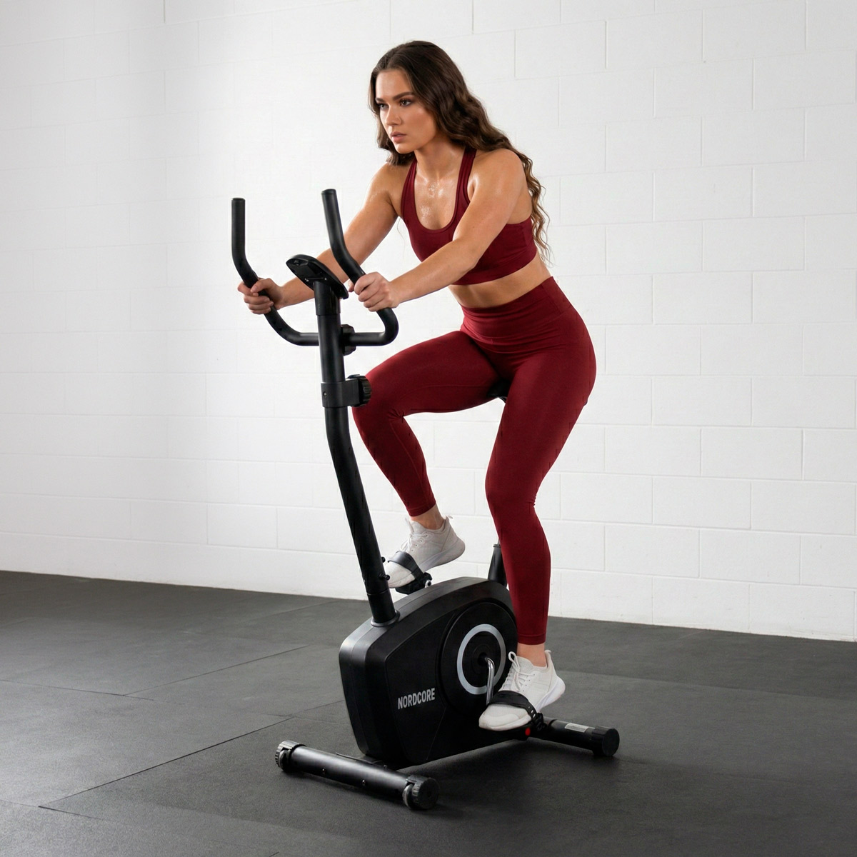 Kobieta w bordowych ubraniach sportowych ćwiczy na czarnym rowerze stacjonarnym przed białą ceglaną ścianą.
