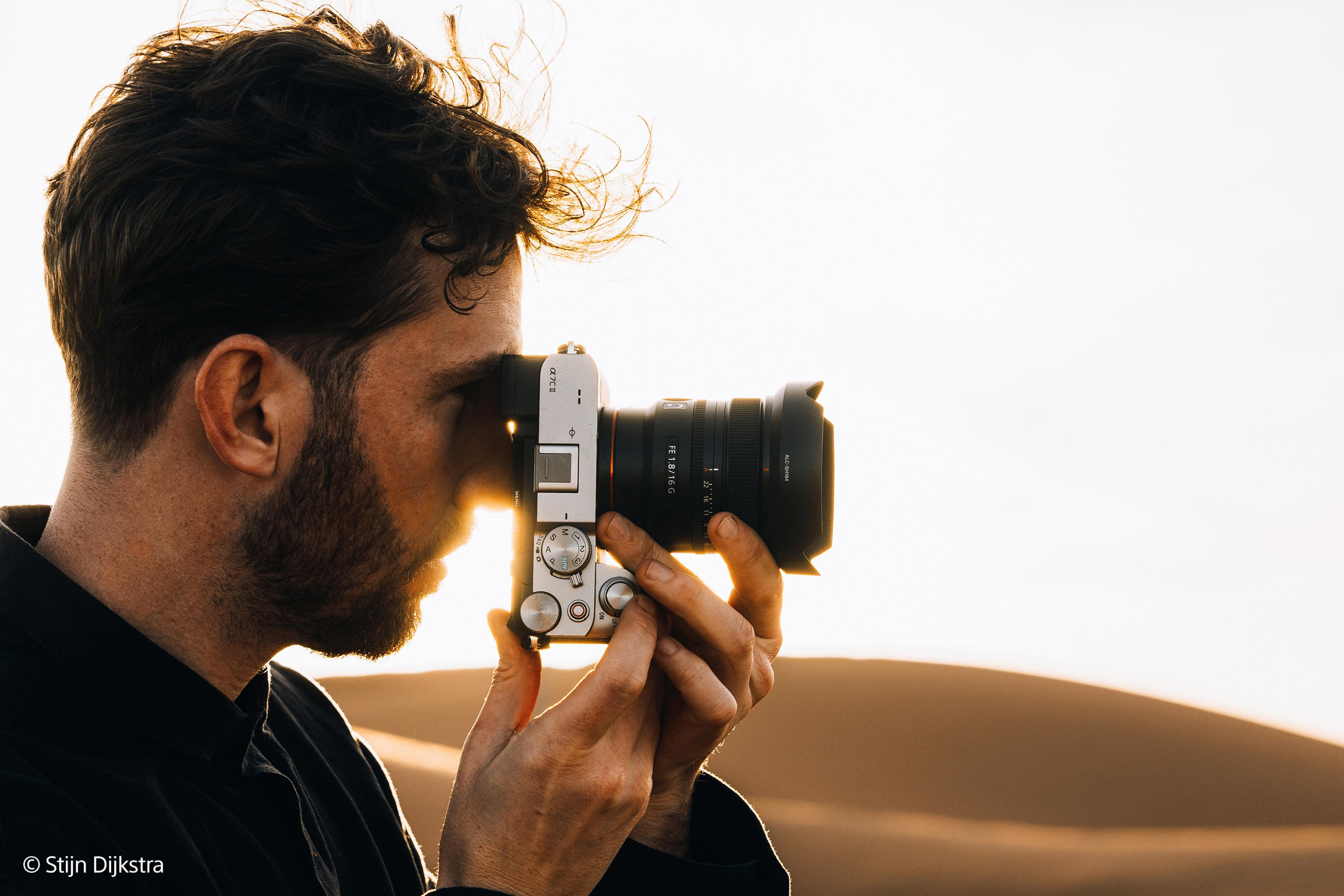 Man in profile photographing with a camera against the sun, sand dunes in background.