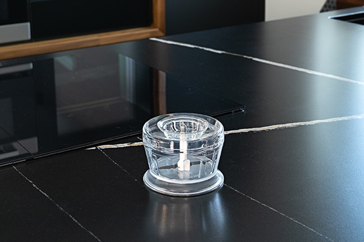 Ein transparenter Mini-Zerkleinerer mit Klingen steht auf einer schwarzen Arbeitsplatte.
