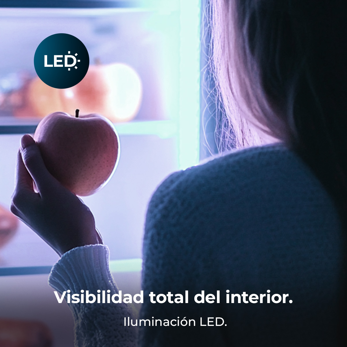 Una persona sostiene una manzana frente a un refrigerador iluminado.