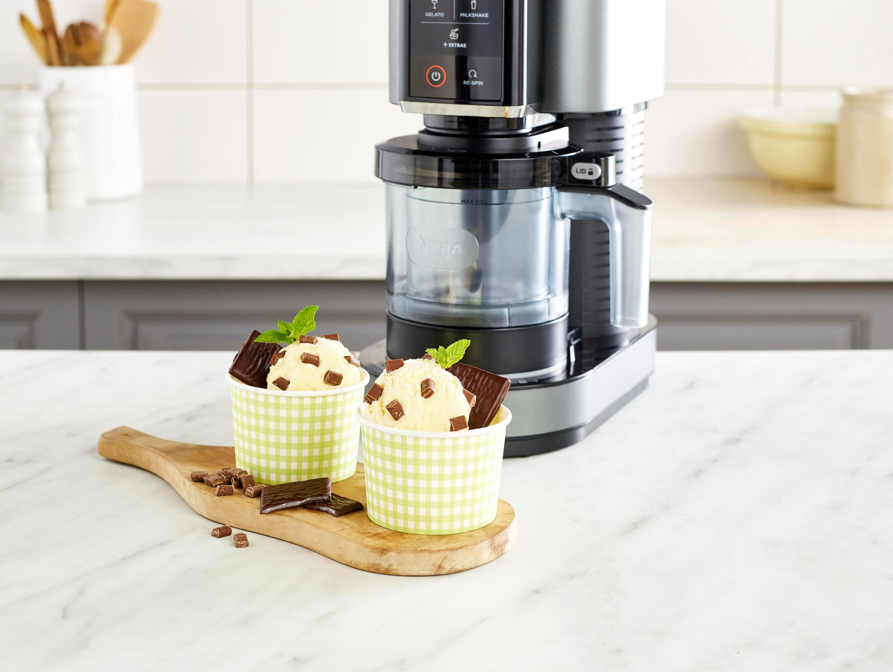 Two ice cream cups with chocolate bits and mint sit on a wooden board next to an ice cream maker.
