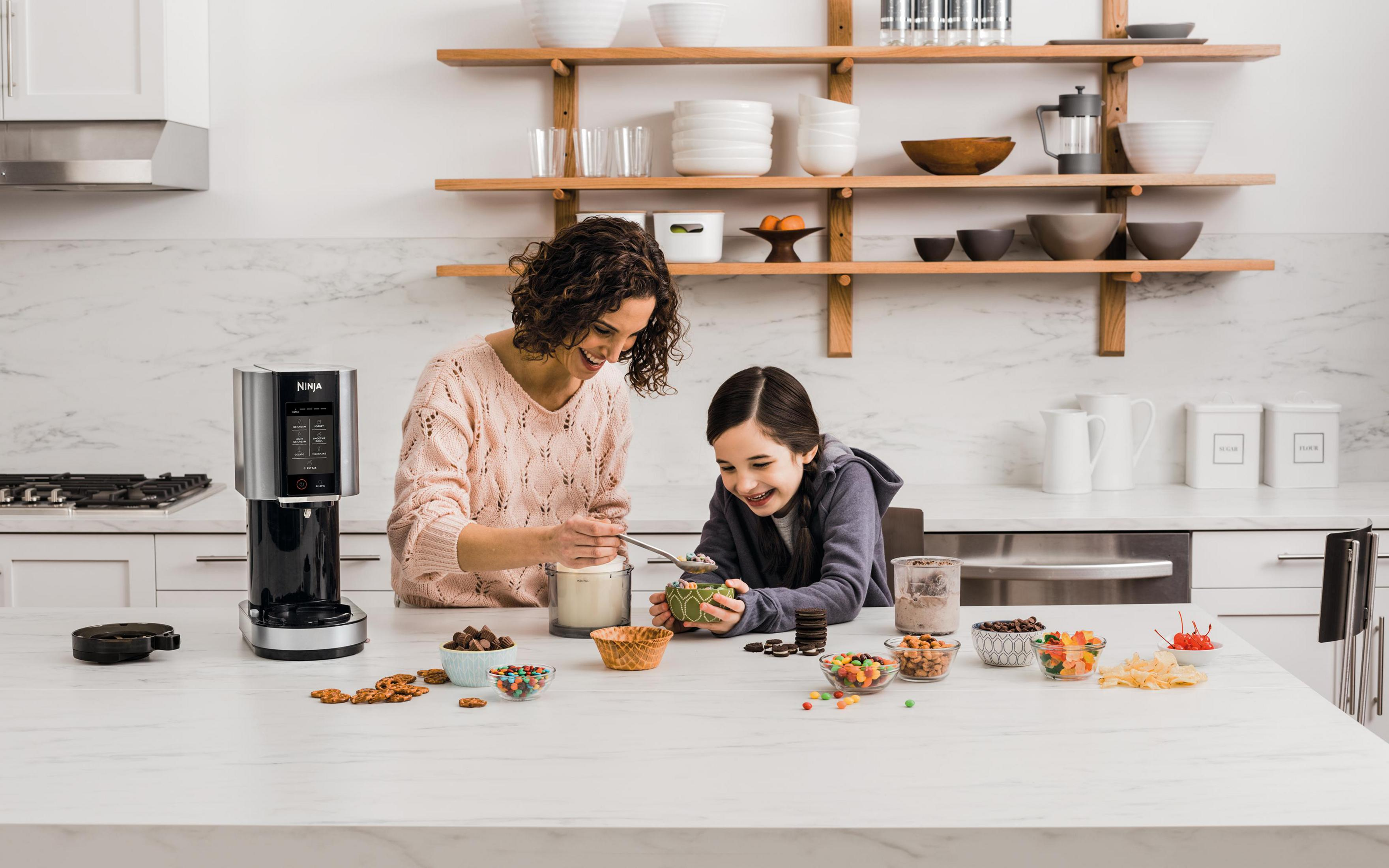 Woman and child make ice cream in a kitchen, with toppings on the counter.