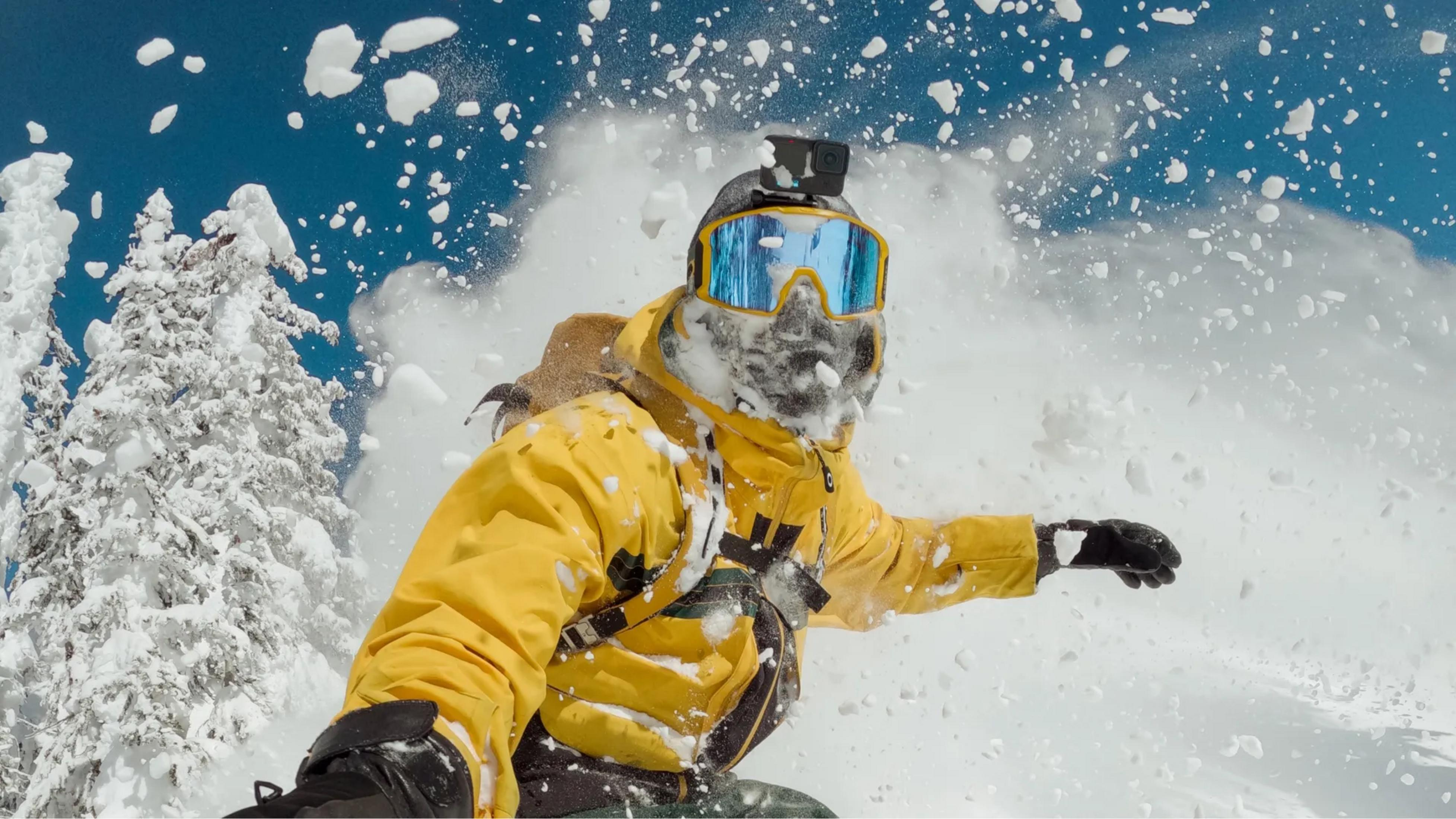 Snowboarder w żółci jedzie przez śnieg, GoPro na kasku, niebieskie gogle.
