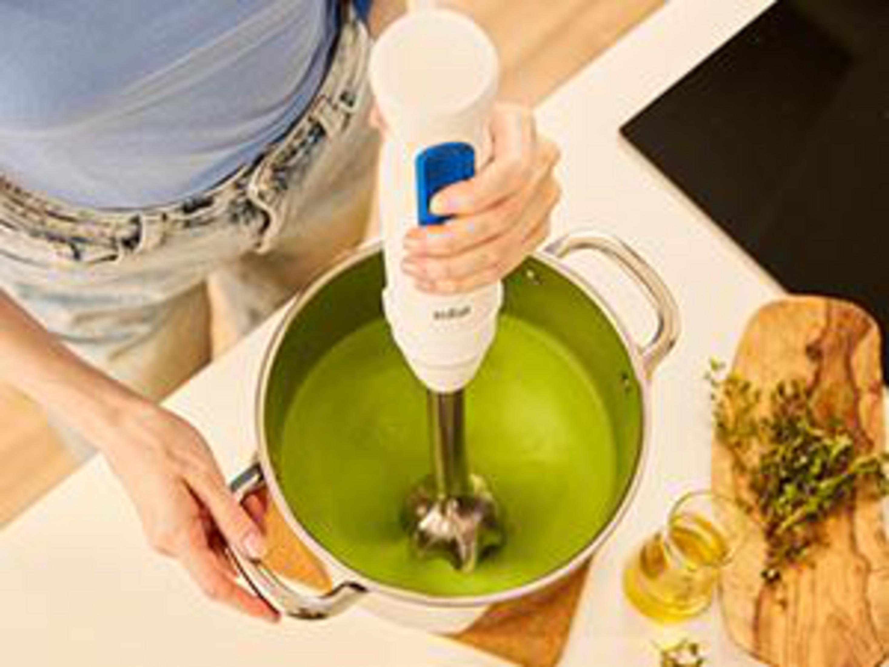 Een hand houdt een staafmixer vast in een pot groene soep.