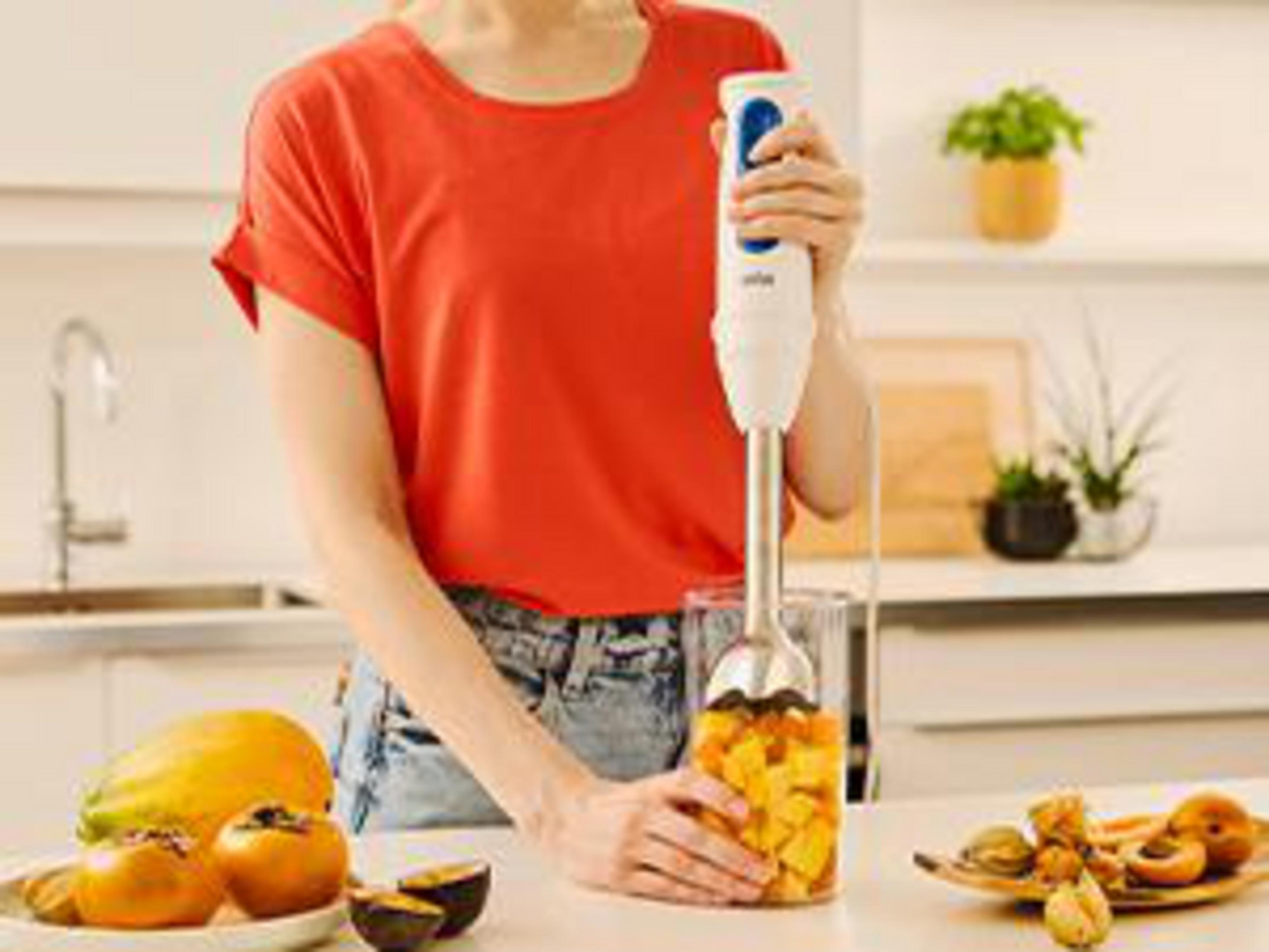 Persoon mixt fruit in een glas met staafmixer in keuken.