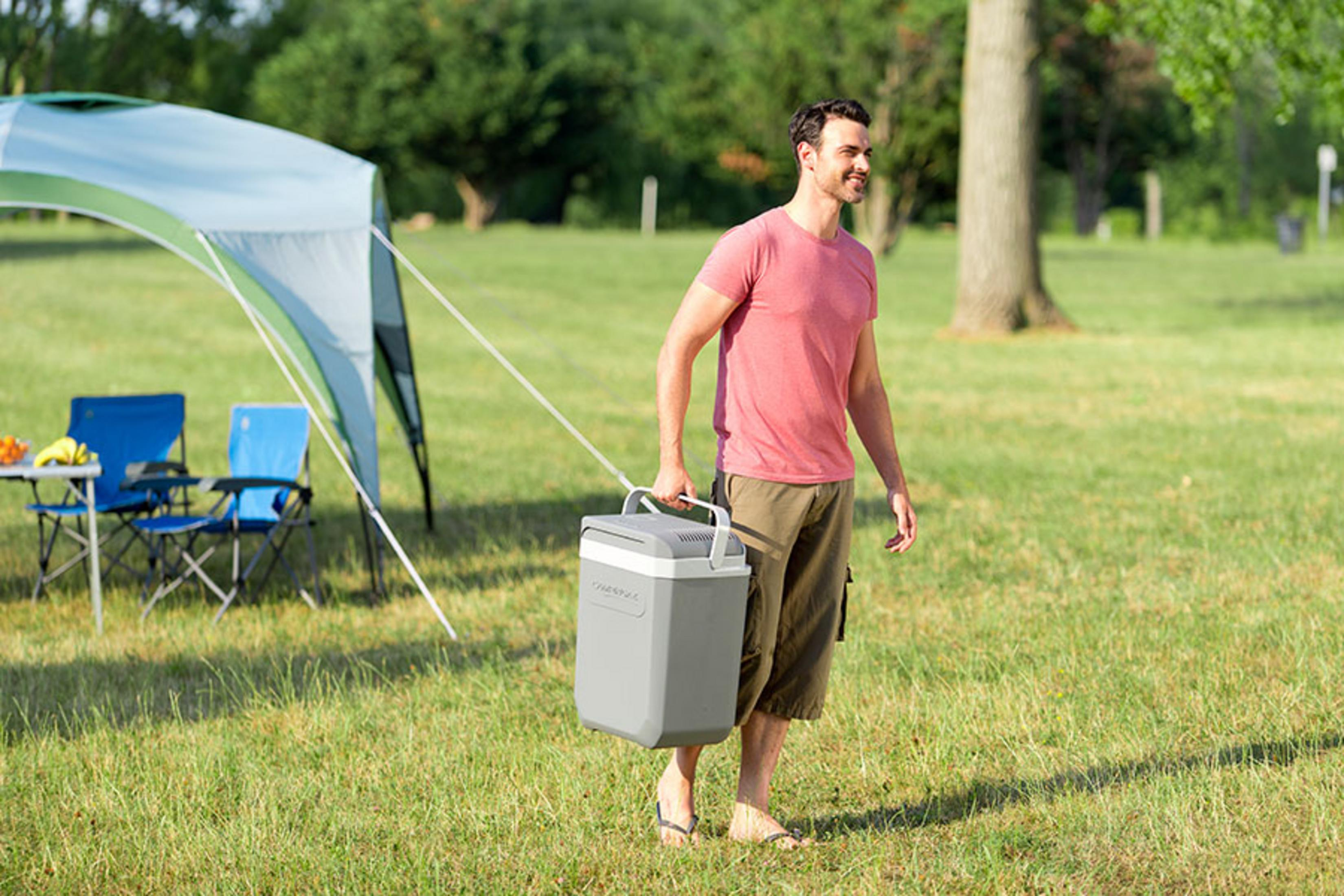 Mężczyzna niesie lodówkę turystyczną na trawie z namiotem.
