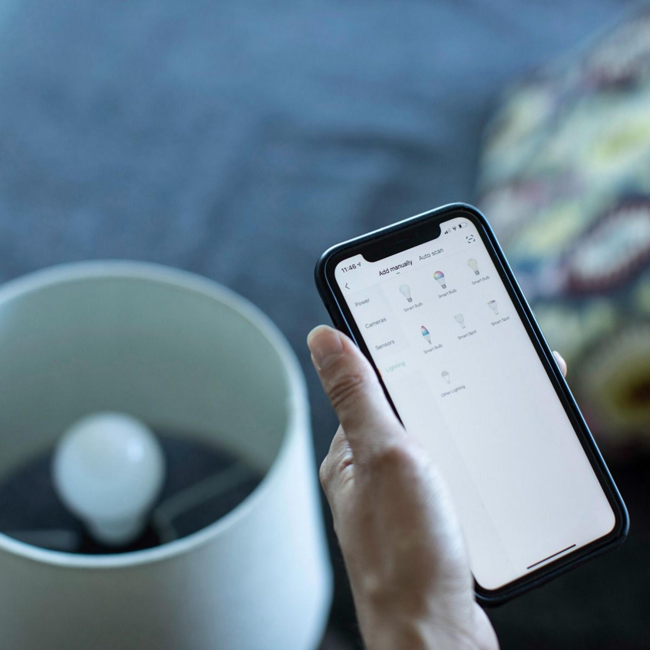 Una mano tiene uno smartphone che mostra un'app per la casa intelligente accanto a una lampada.