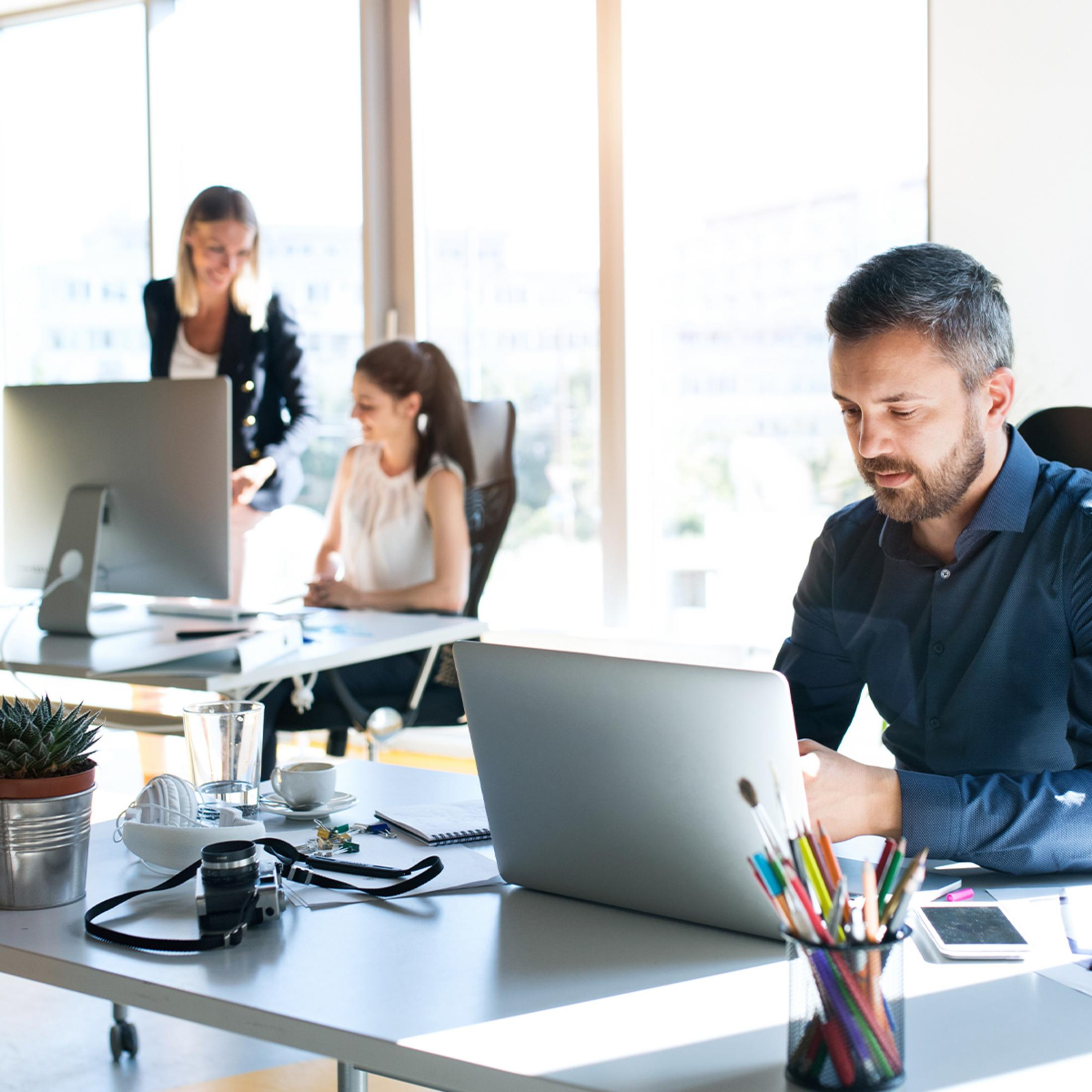 Männer und Frauen arbeiten in Büros vor Computern. Buntstifte liegen auf einem Schreibtisch.