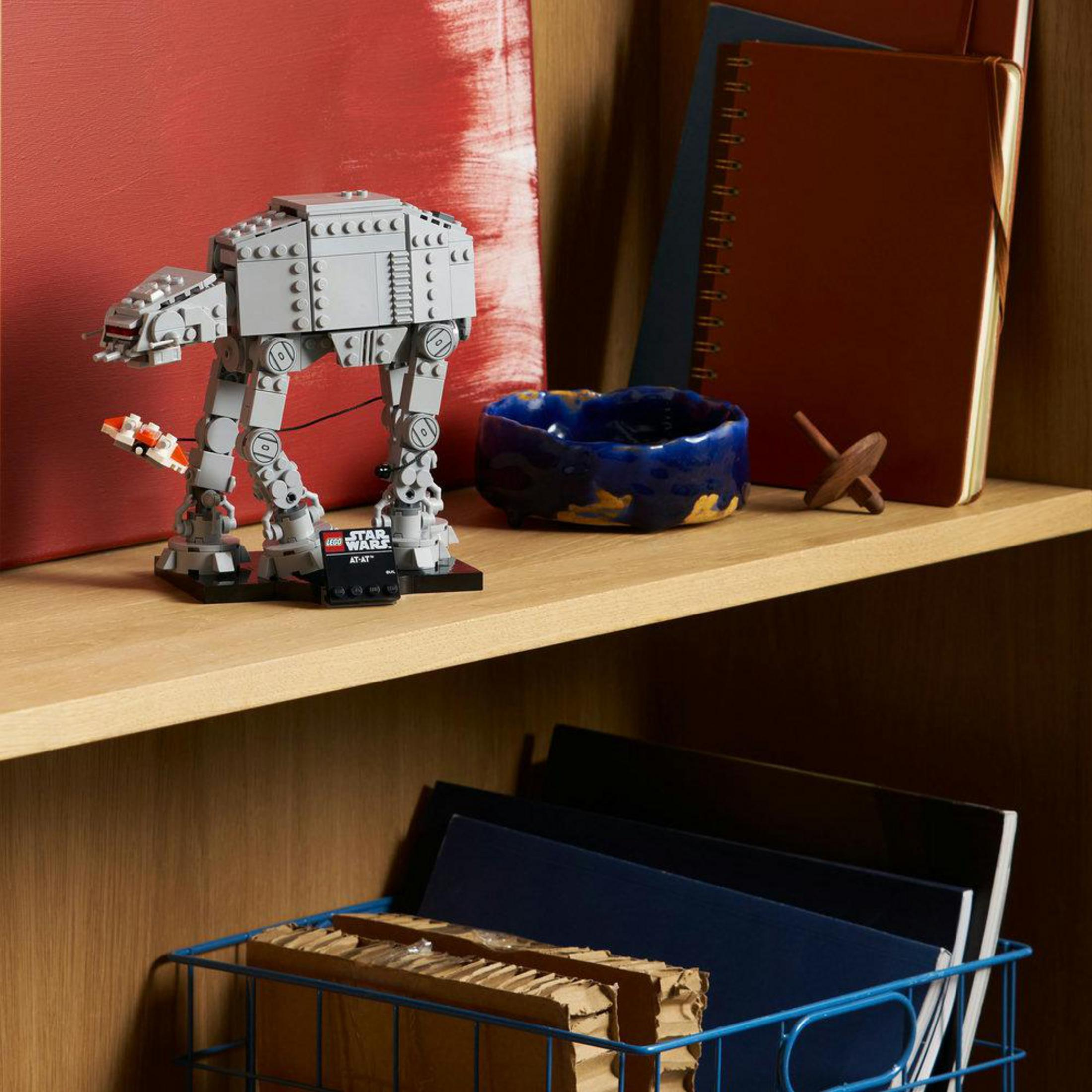 Gri Lego AT-AT modeli, mavi seramik bir kabın yanında rafta duruyor.