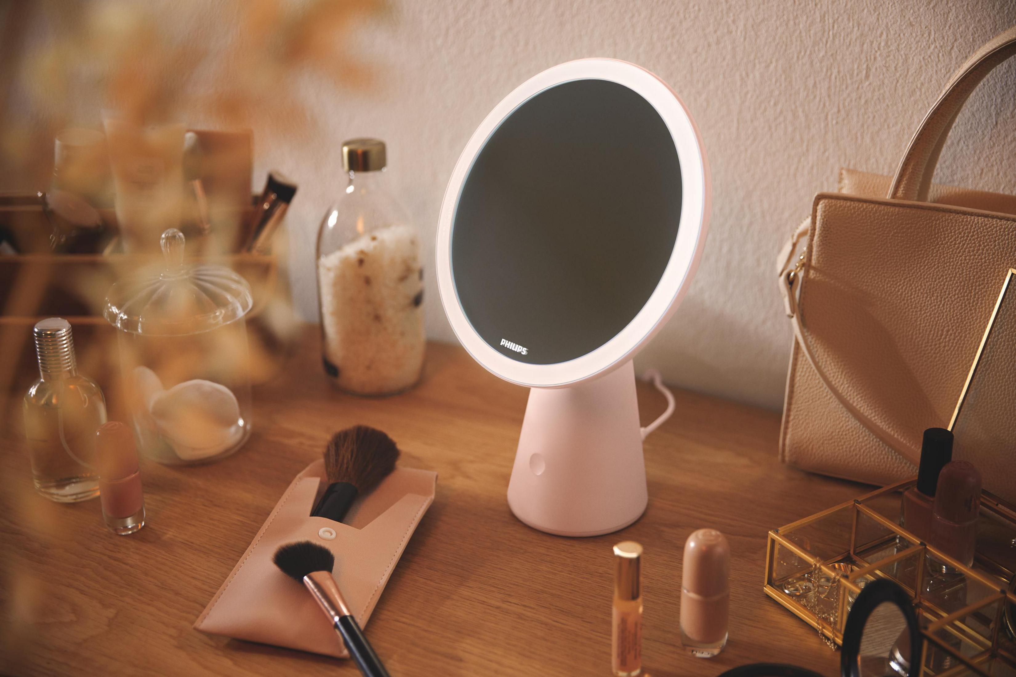 Ein rosa Schminkspiegel mit Licht steht auf einem Holztisch neben Kosmetik.